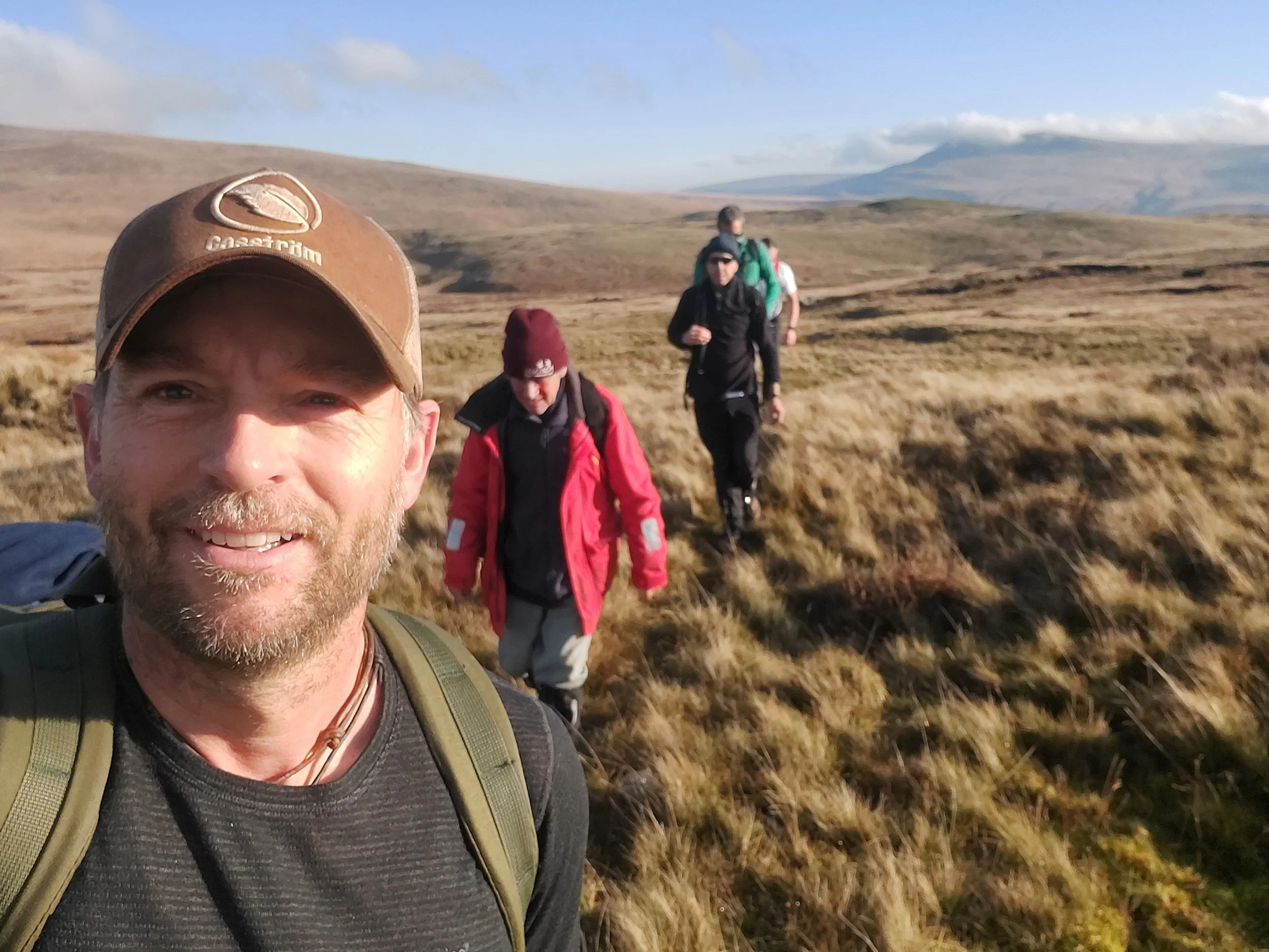 A group of four hikers walking through a grassy landscape with hills and mountains in the background, one man leading and smiling at the camera.