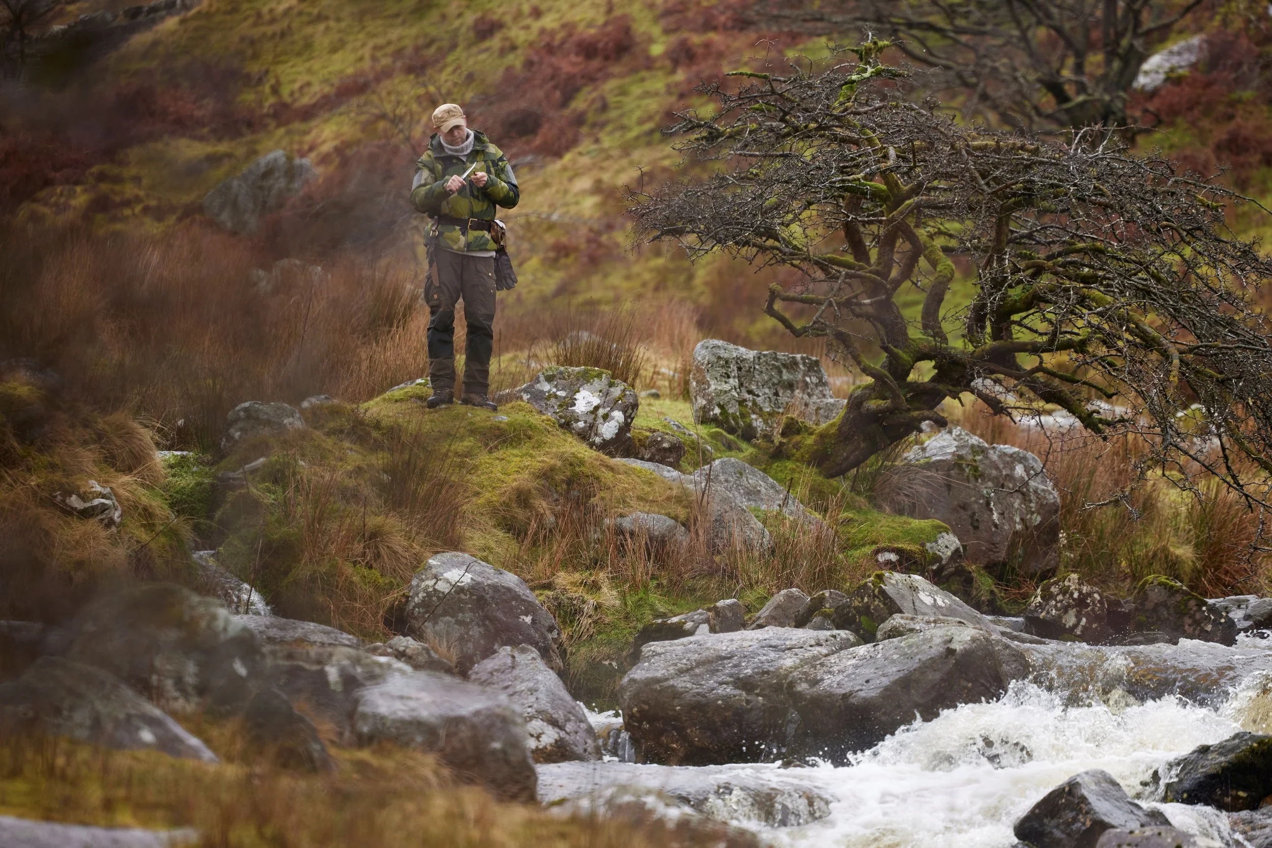 A man dressed in outdoor gear, holding a map or compass, standing on mossy rocks near a stream in a rugged, hilly landscape with leafless tree and brown grass.
