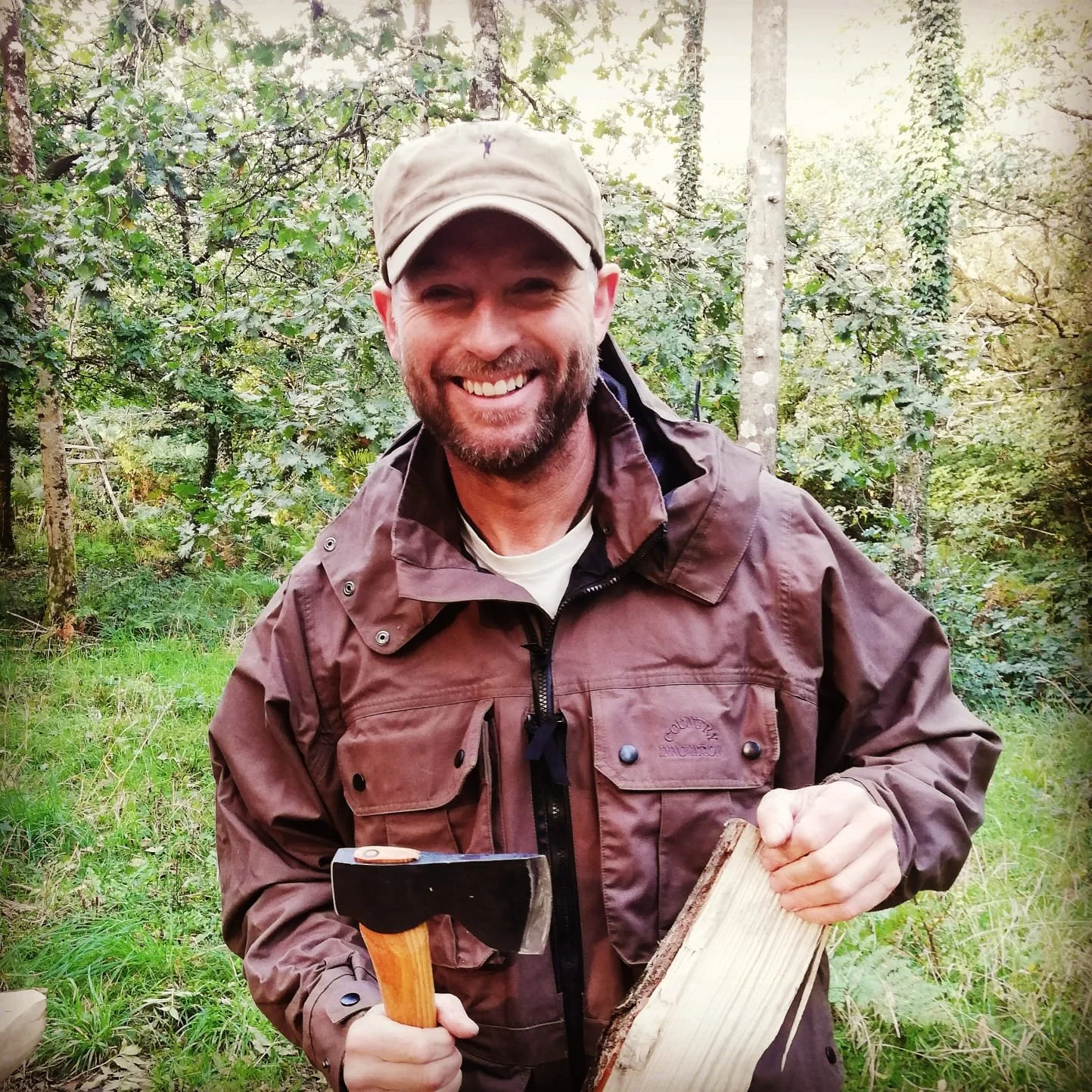 A man with a beard and cap smiling outdoors in a wooded area, holding an axe and a piece of wood.