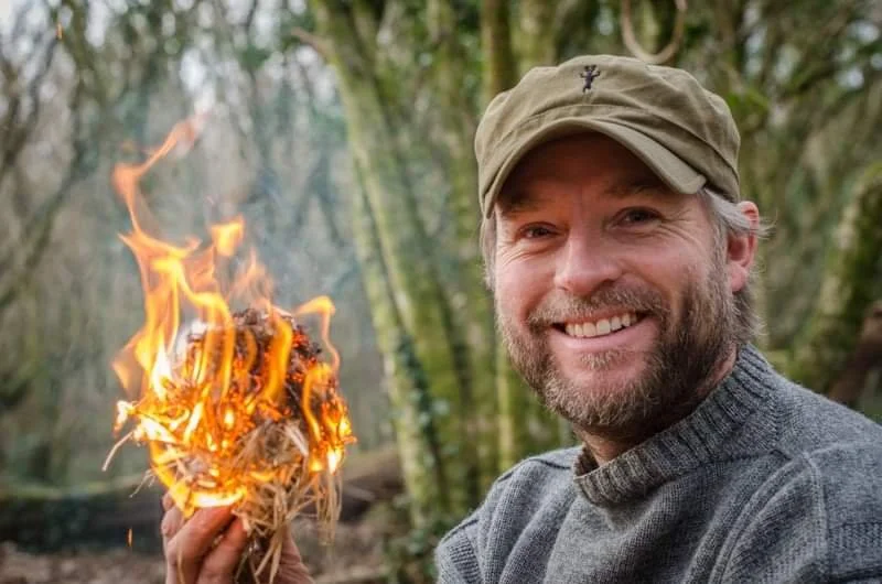 A man outdoors in a forest, smiling and holding a small burning bundle of twigs.