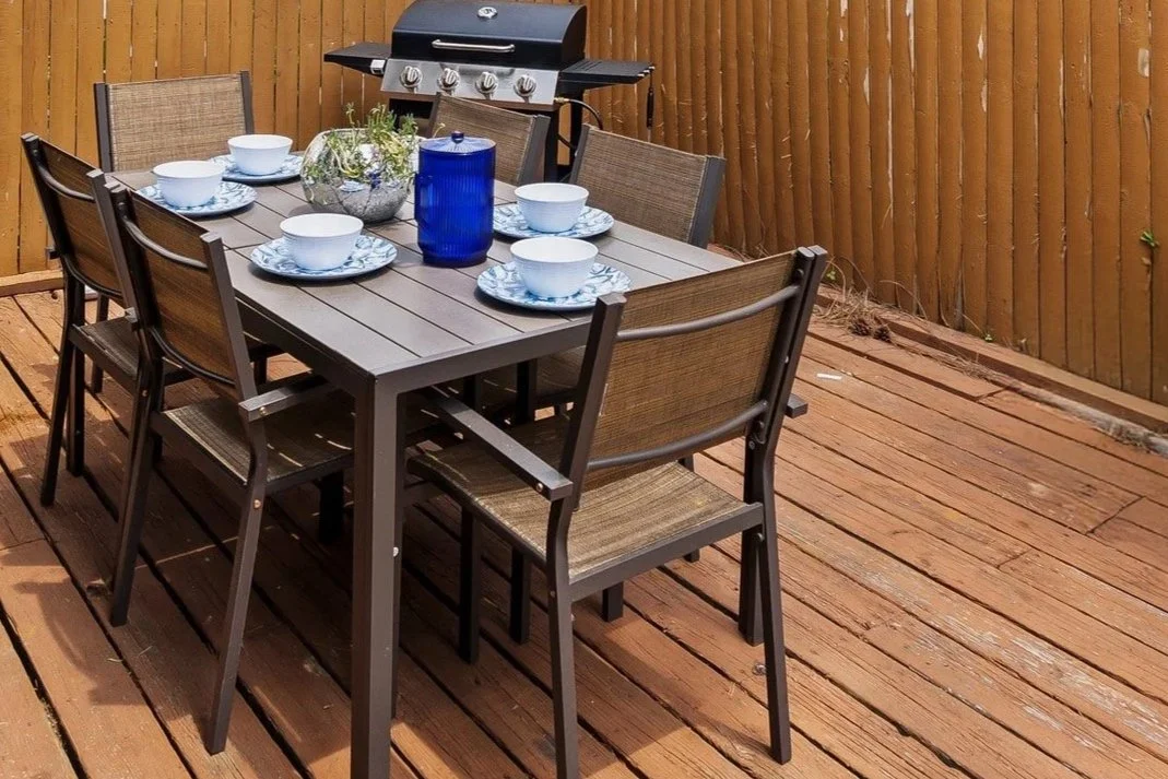 Outdoor dining table with six chairs, set with white bowls and plates, a large blue pitcher, a glass terrarium with green plants, and a black gas grill in a fenced backyard with wooden deck.