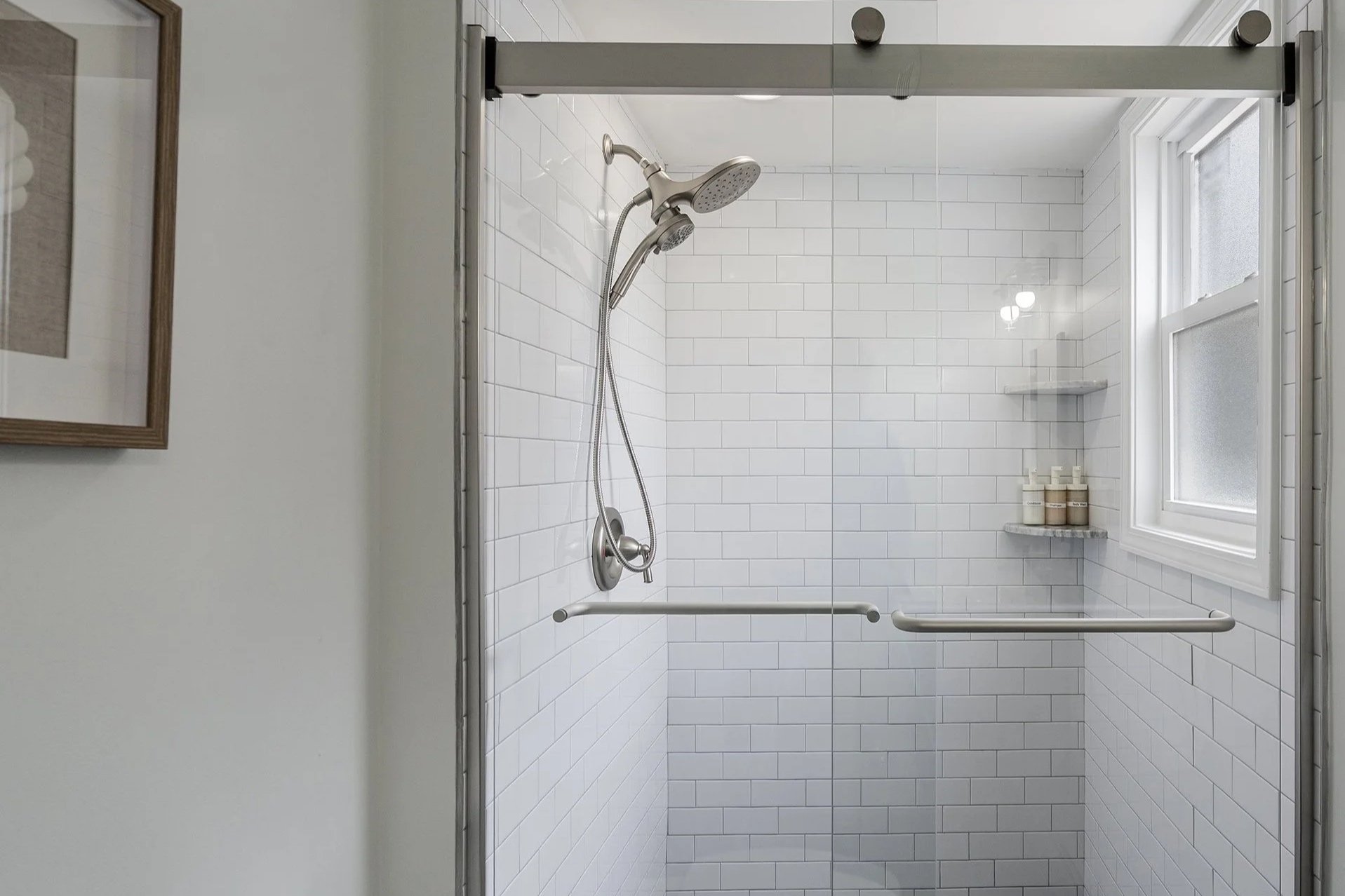 A newly renovated walk-in shower with white subway tiles, a handheld showerhead, a grab bar, and a window with frosted glass in a bathroom.