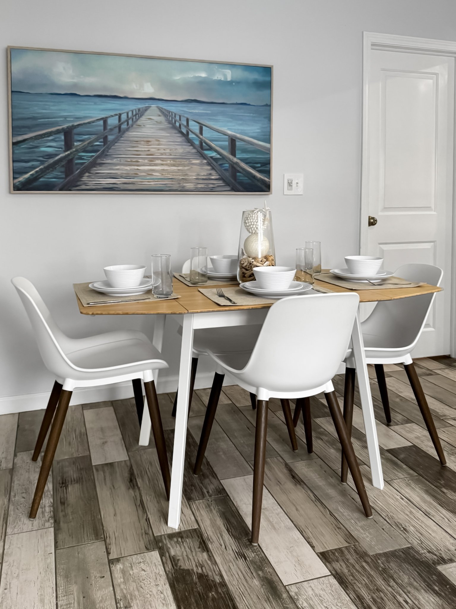 Dining table set for three with white bowls, glasses, and cutlery, decorated with a glass vase filled with seashells and white decorative orbs, placed against a white wall with a large painting of a wooden pier extending over water.