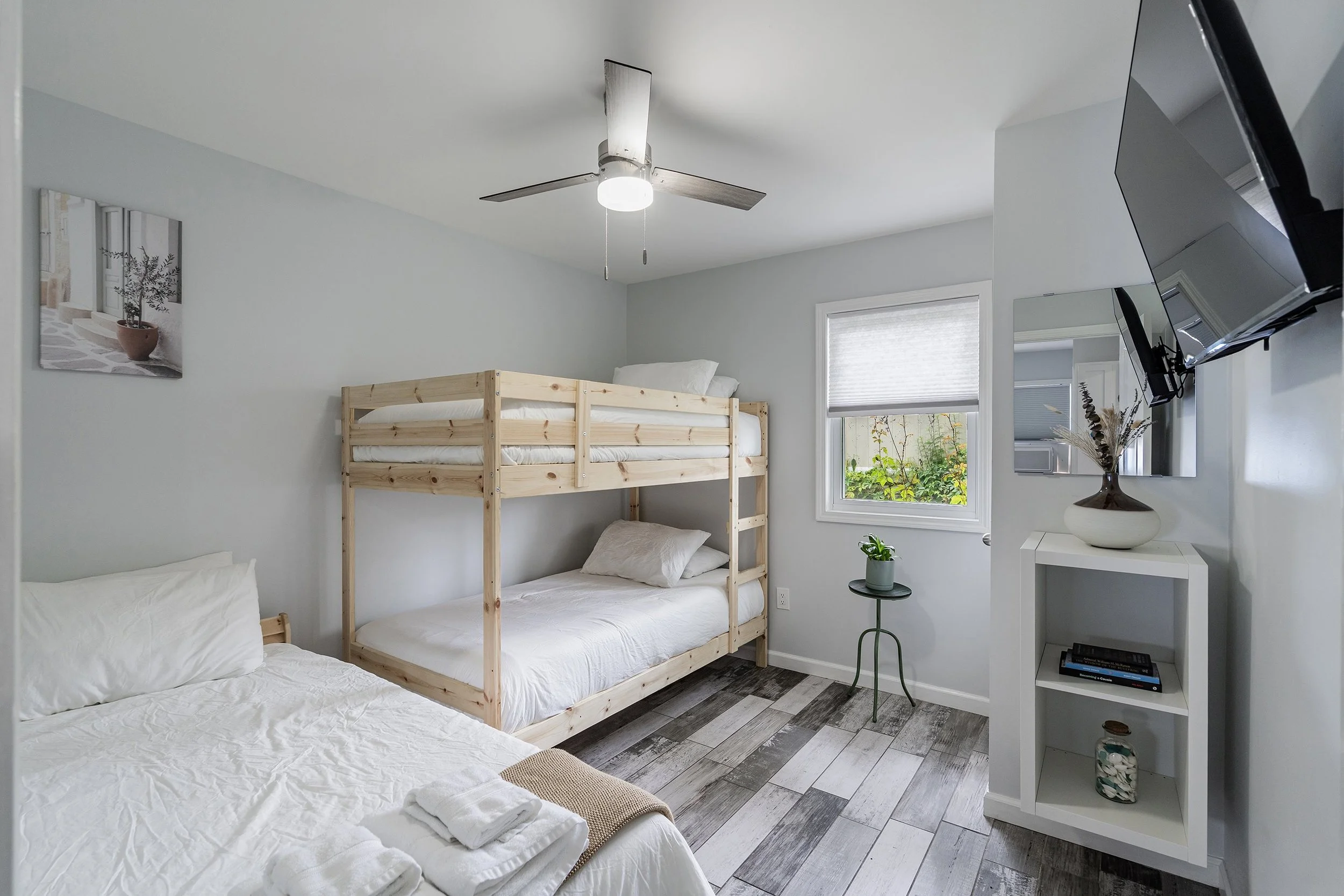 Cozy bedroom with two beds, including a wooden bunk bed and a double bed, in a room with white walls, a ceiling fan, a small window with a roll-up shade, a small side table with a potted plant, and a wall-mounted TV.