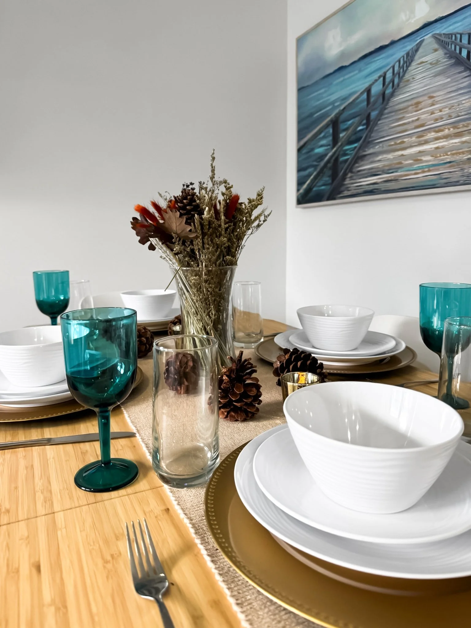 A dining table set with white porcelain dishes, gold-rimmed chargers, colorful glassware, and a centerpiece of dried flowers and pinecones, with a framed seascape painting on the wall.
