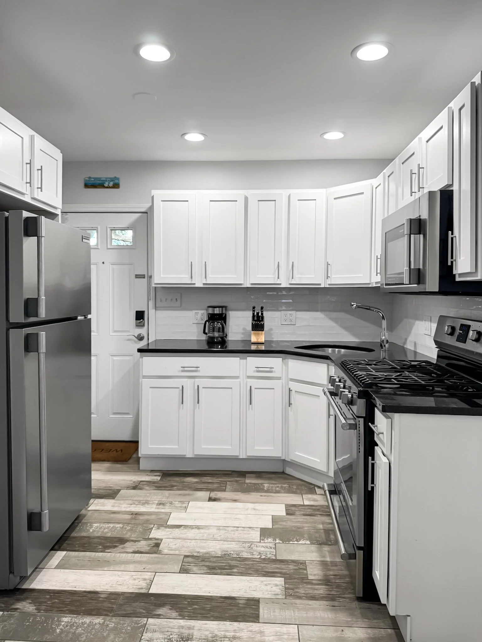 A modern kitchen with white cabinets, stainless steel appliances including a refrigerator and microwave, a black countertop, a coffee maker, tea kettle, BBQ set.