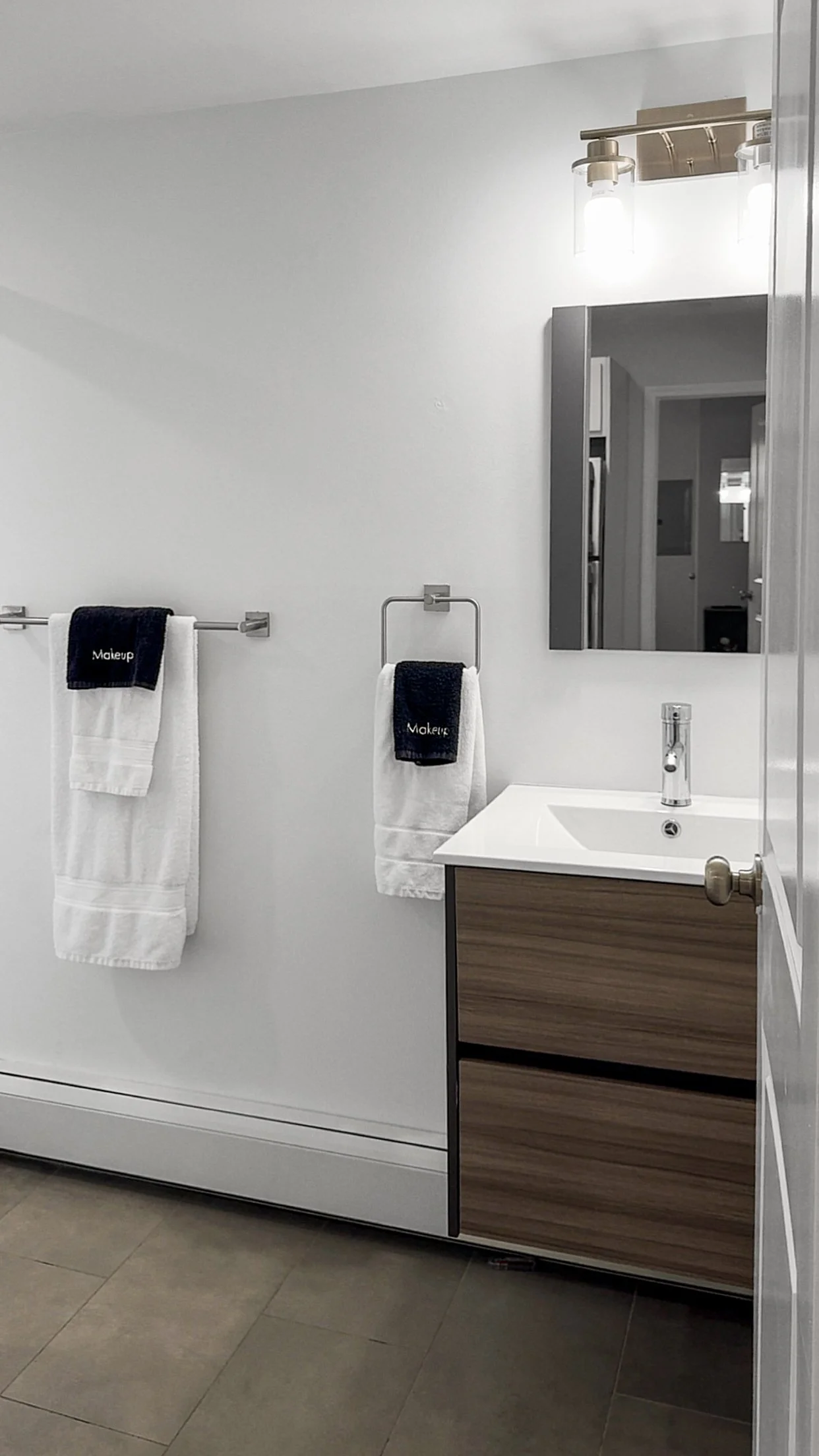 A bathroom with a white wall, a mirror, a wooden cabinet with a white sink, and three towels with the word "Makeup" embroidered on them, hanging on a towel bar and a towel ring.