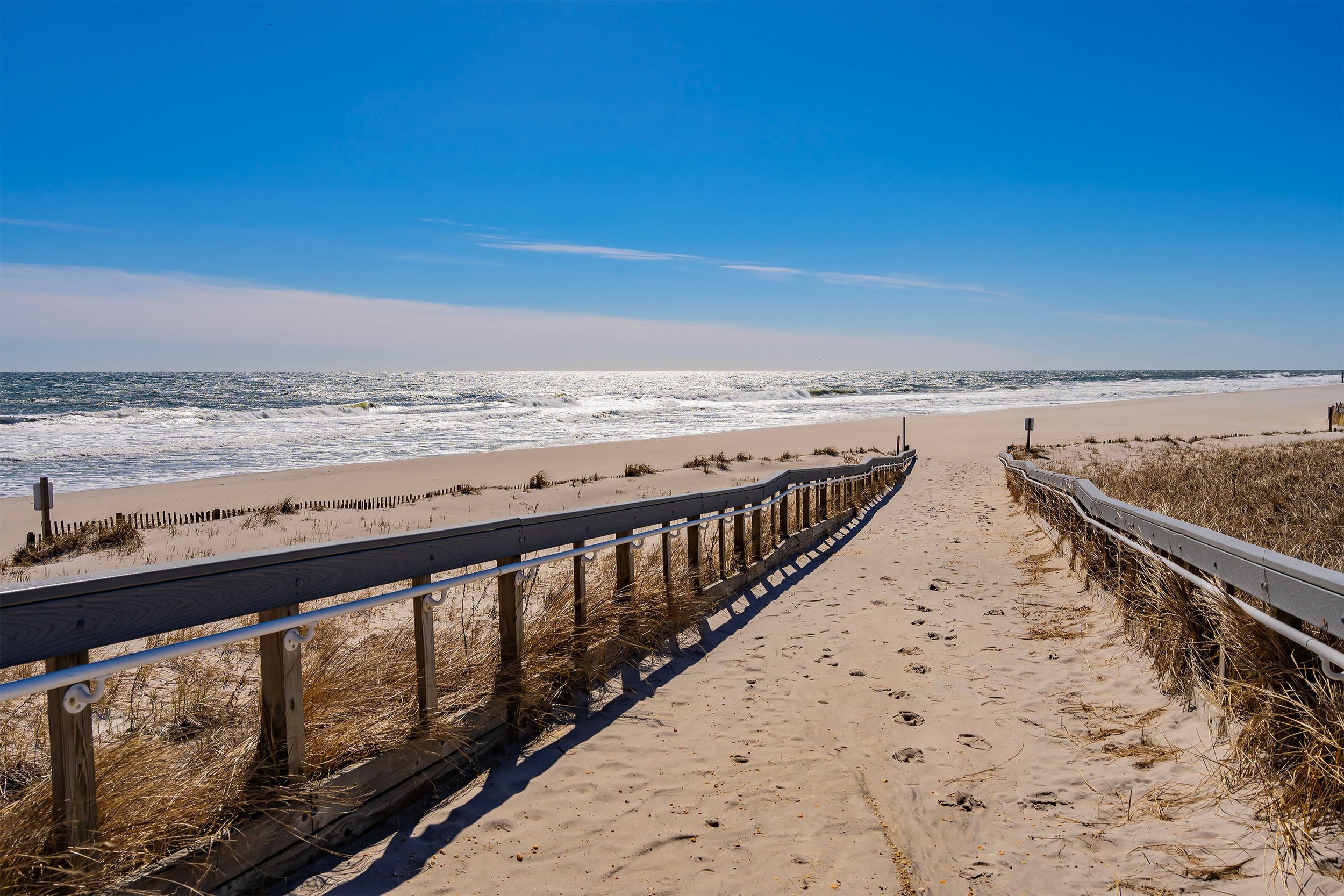 Pathway leading to a sandy beach with a view of the ocean, blue sky with some clouds, and grasses on the sides.