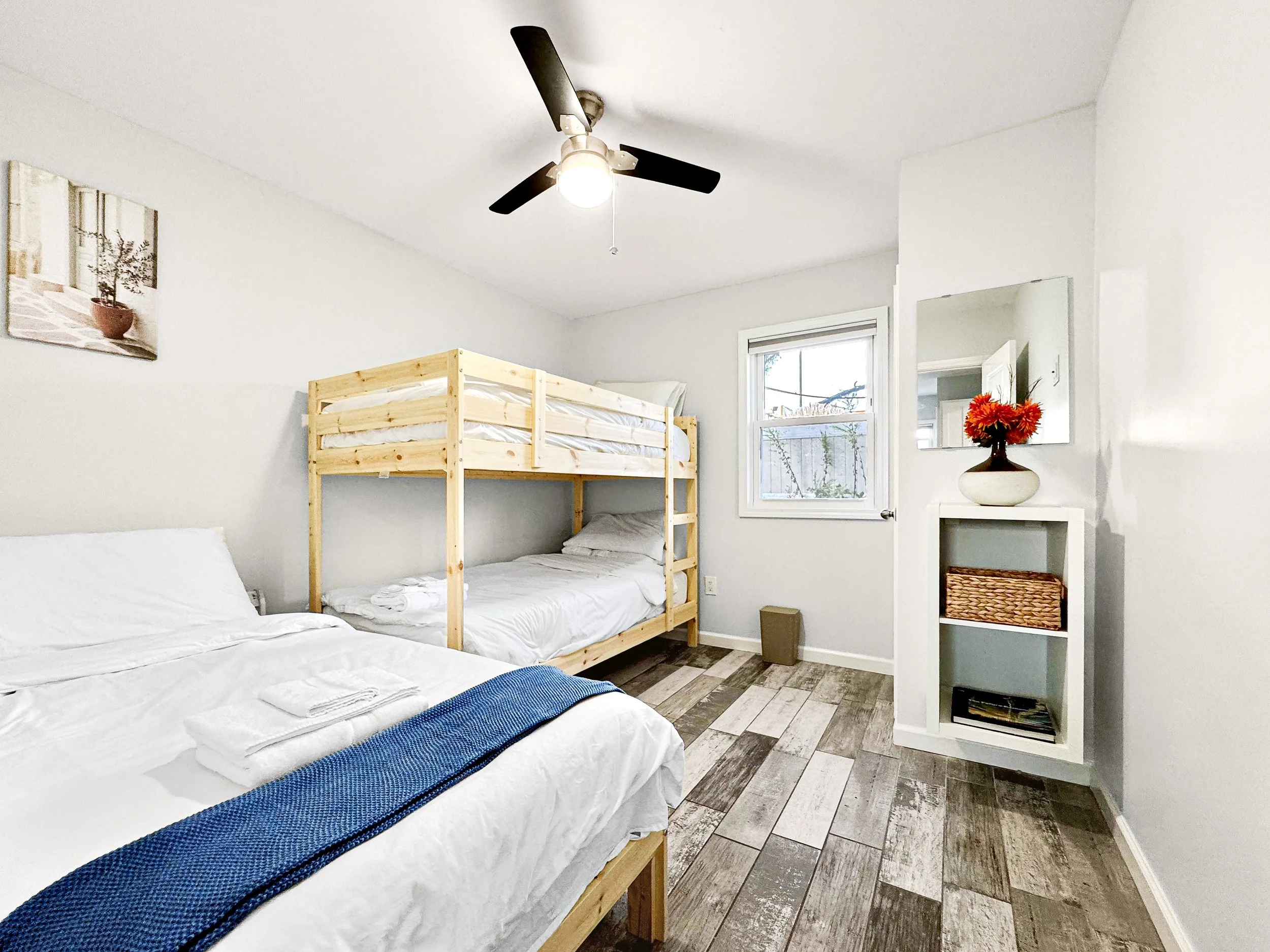Secondary bedroom with two beds, one twin and one bunk bed, white linens, ceiling fan, a wall-mounted TV,  small white shelf with decor, and small round table with decor.