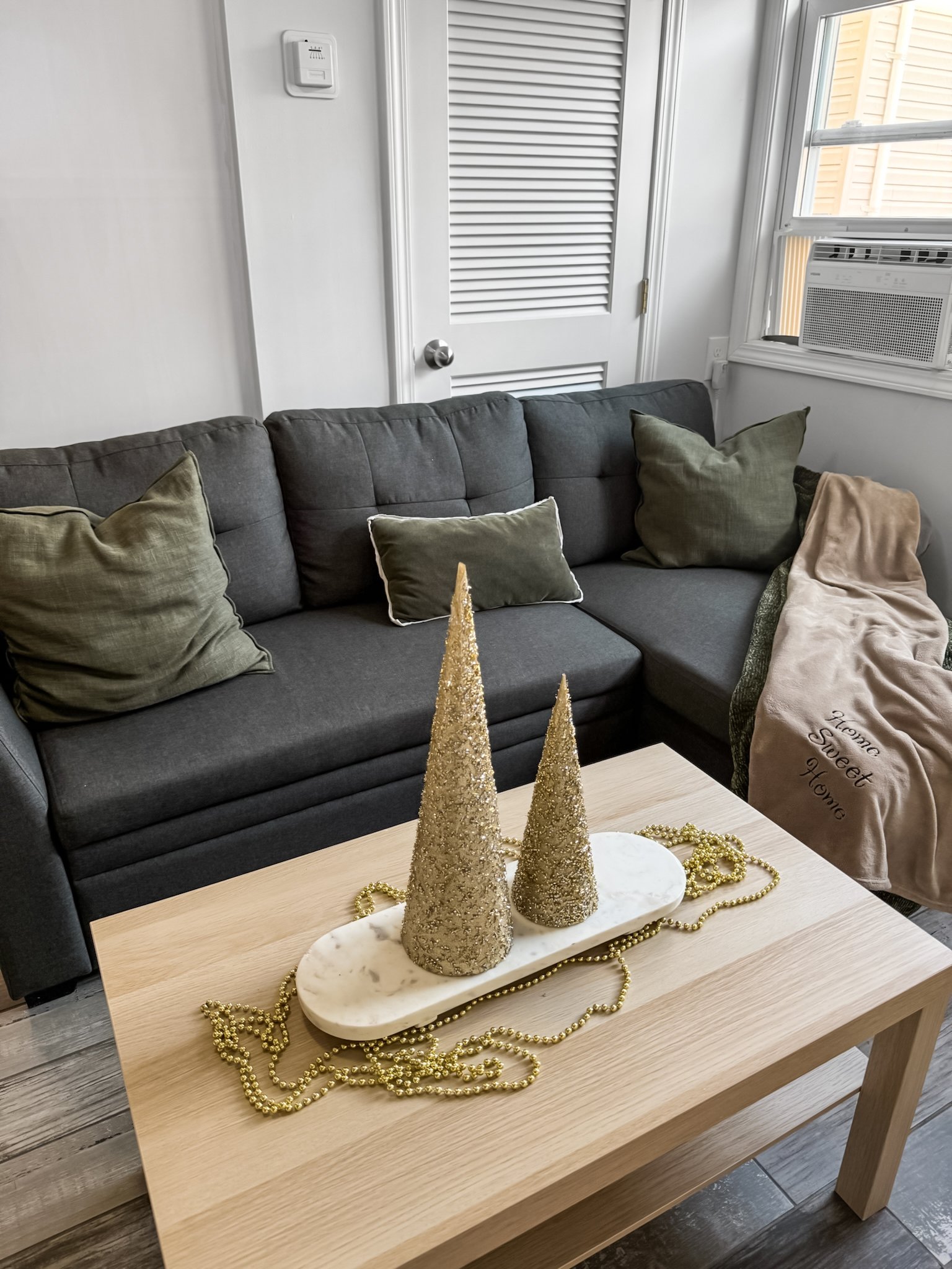Decorative Christmas centerpiece with two glittery gold cone-shaped trees and gold bead garland on a white marble tray, positioned on a light wooden coffee table in front of a dark gray sofa with green pillows.