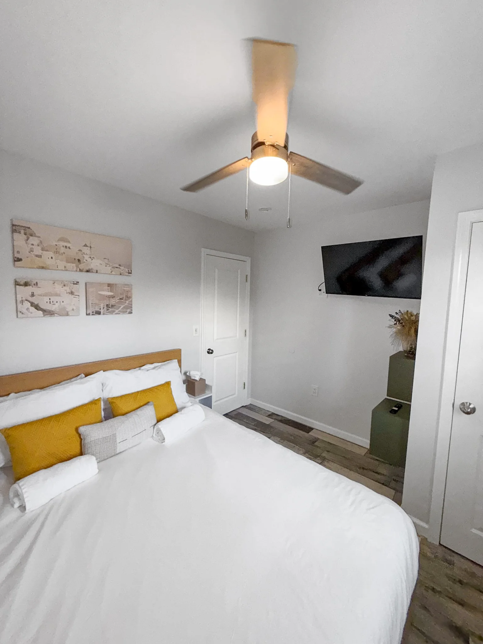A bedroom with a Queen bed, fresh linen, wall art, a ceiling fan, and a mounted flat-screen TV.