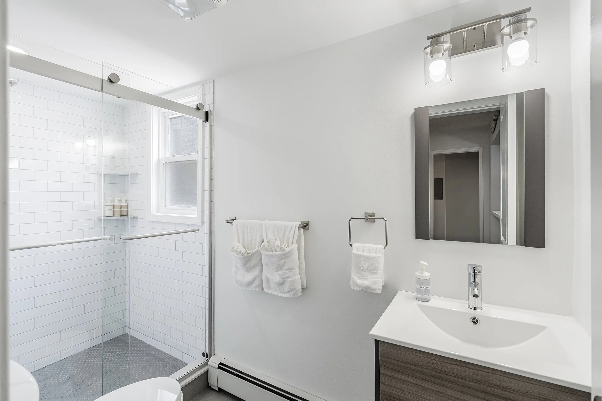 Modern bathroom with walk-in shower, white subway tiles, window, white walls, towel rack with towels, mirror, sink, and soap dispenser.