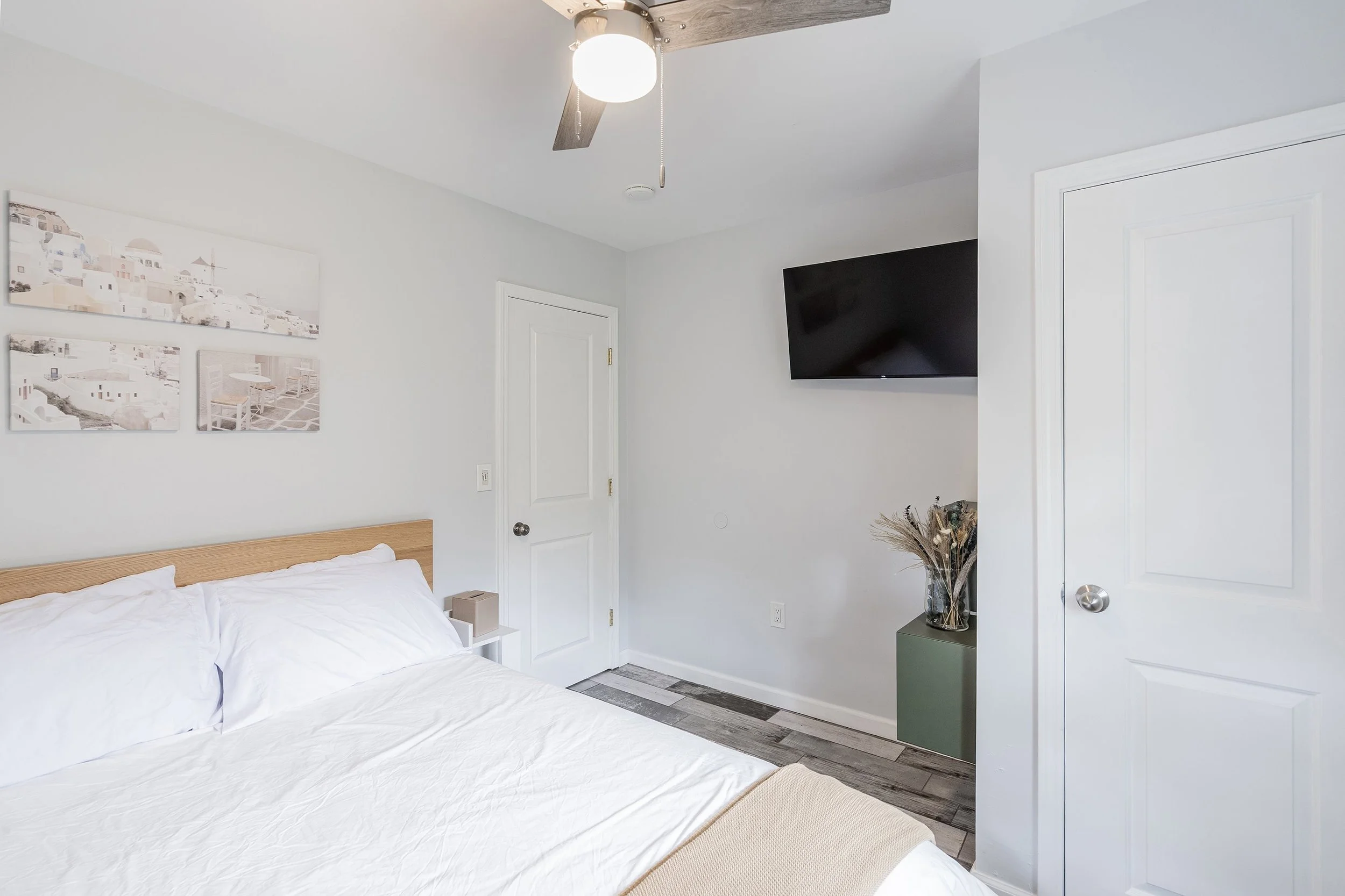 A bedroom with a white bed, wooden headboard, wall art, a wall-mounted TV, a ceiling fan, and a door.