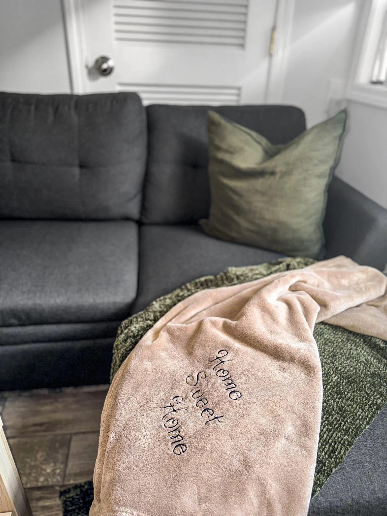 A dark gray couch with a green pillow and a beige throw blanket embroidered with 'Home Sweet Home,' in a living room with a white door and window.