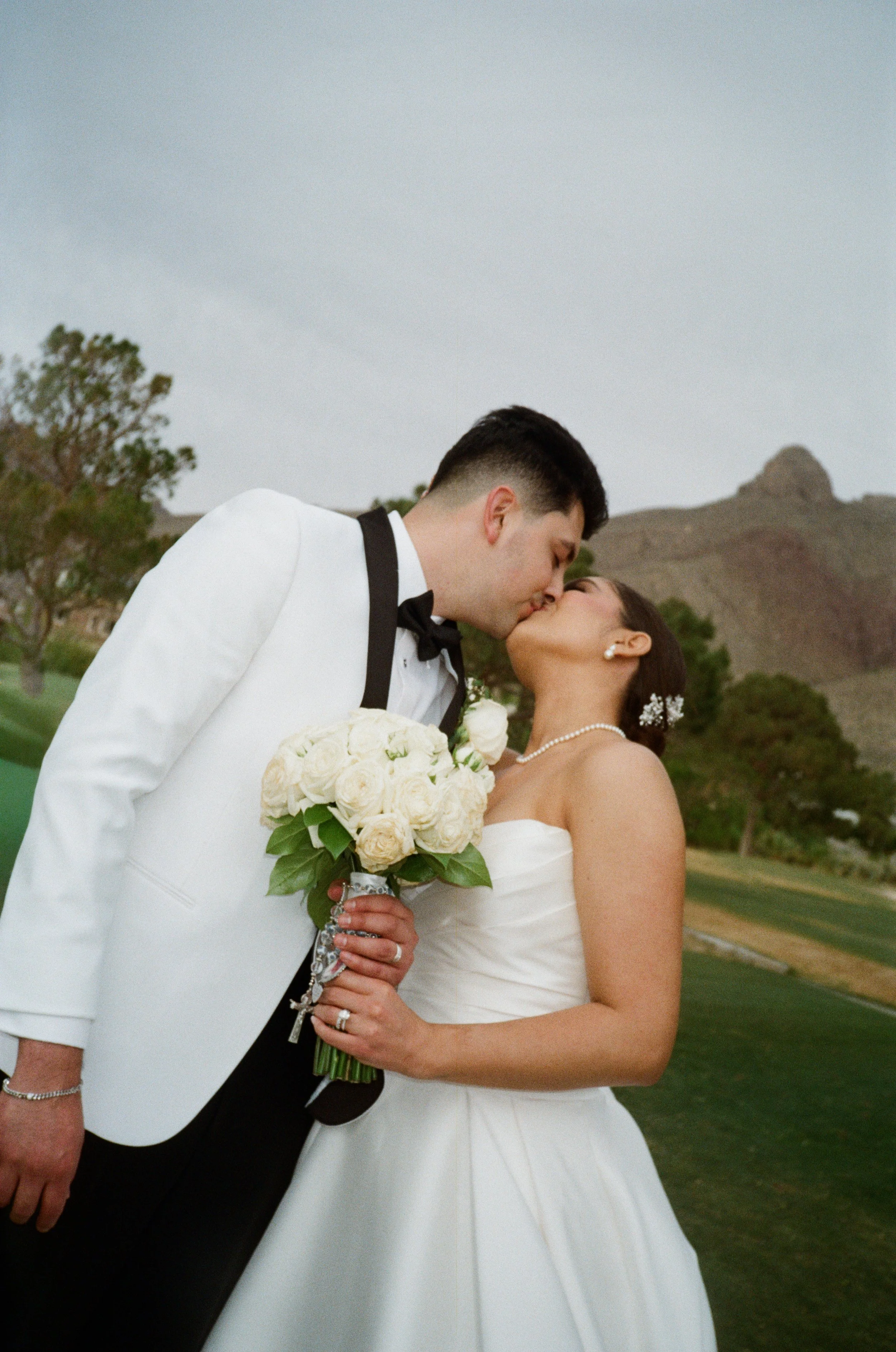 A bride and groom kiss during their wedding outdoors, with the groom in a white tuxedo and the bride in a strapless white wedding dress holding a bouquet of white roses. The background features trees and hills.