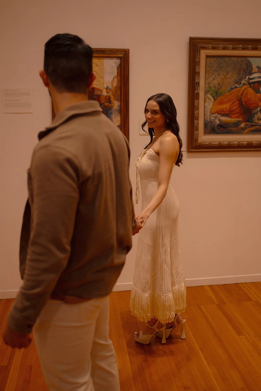 A woman in a beige dress holding hands with a man in a tan jacket, standing in an art gallery. The woman is smiling, and there are framed paintings on the wall behind them.
