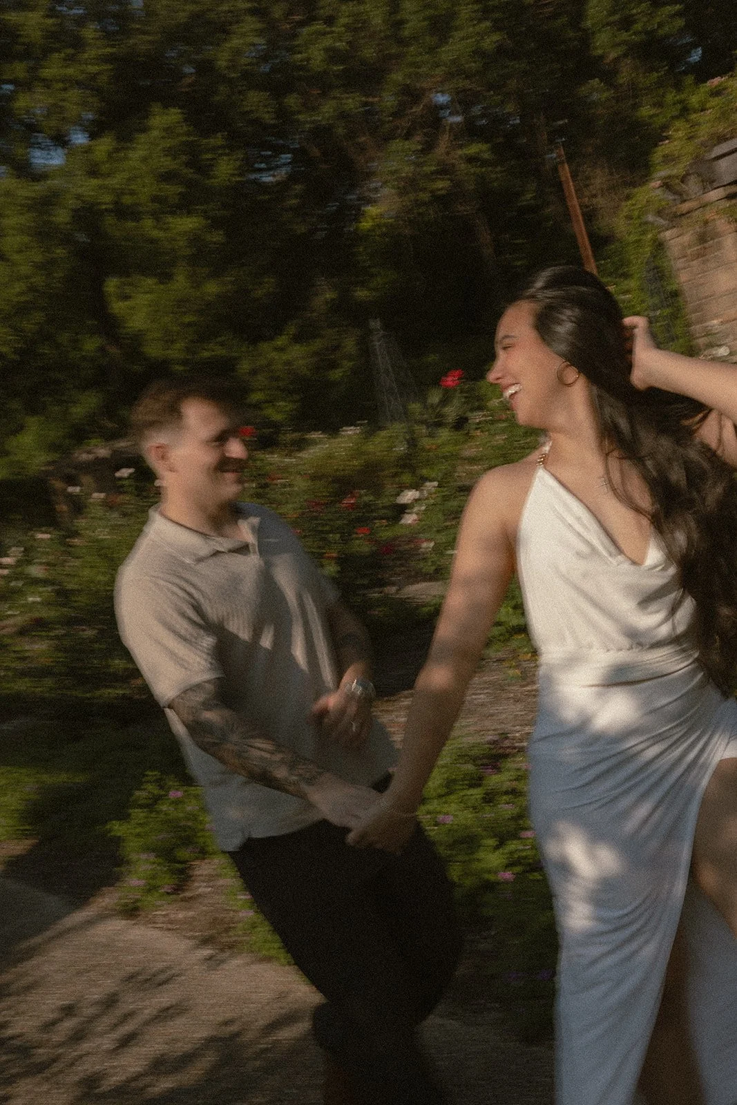 A young man and woman holding hands and smiling, dancing outdoors at night with trees and flowers in the background.