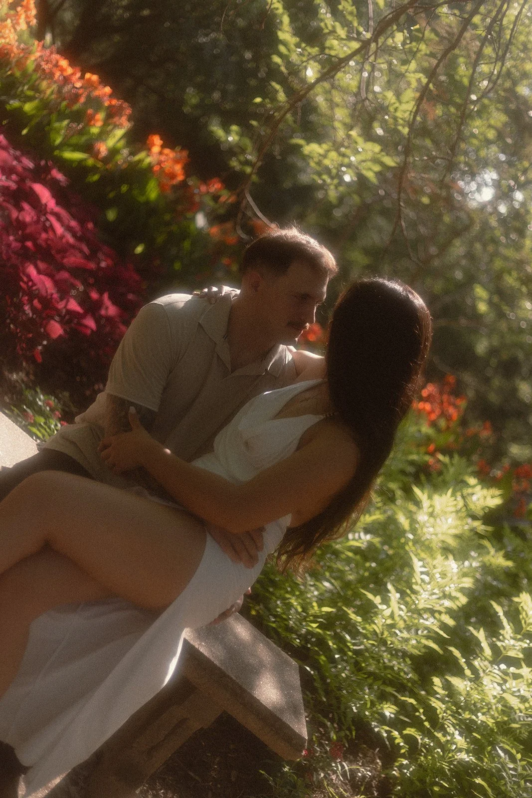 A man and woman sitting closely together outdoors surrounded by colorful foliage, sharing an intimate moment.