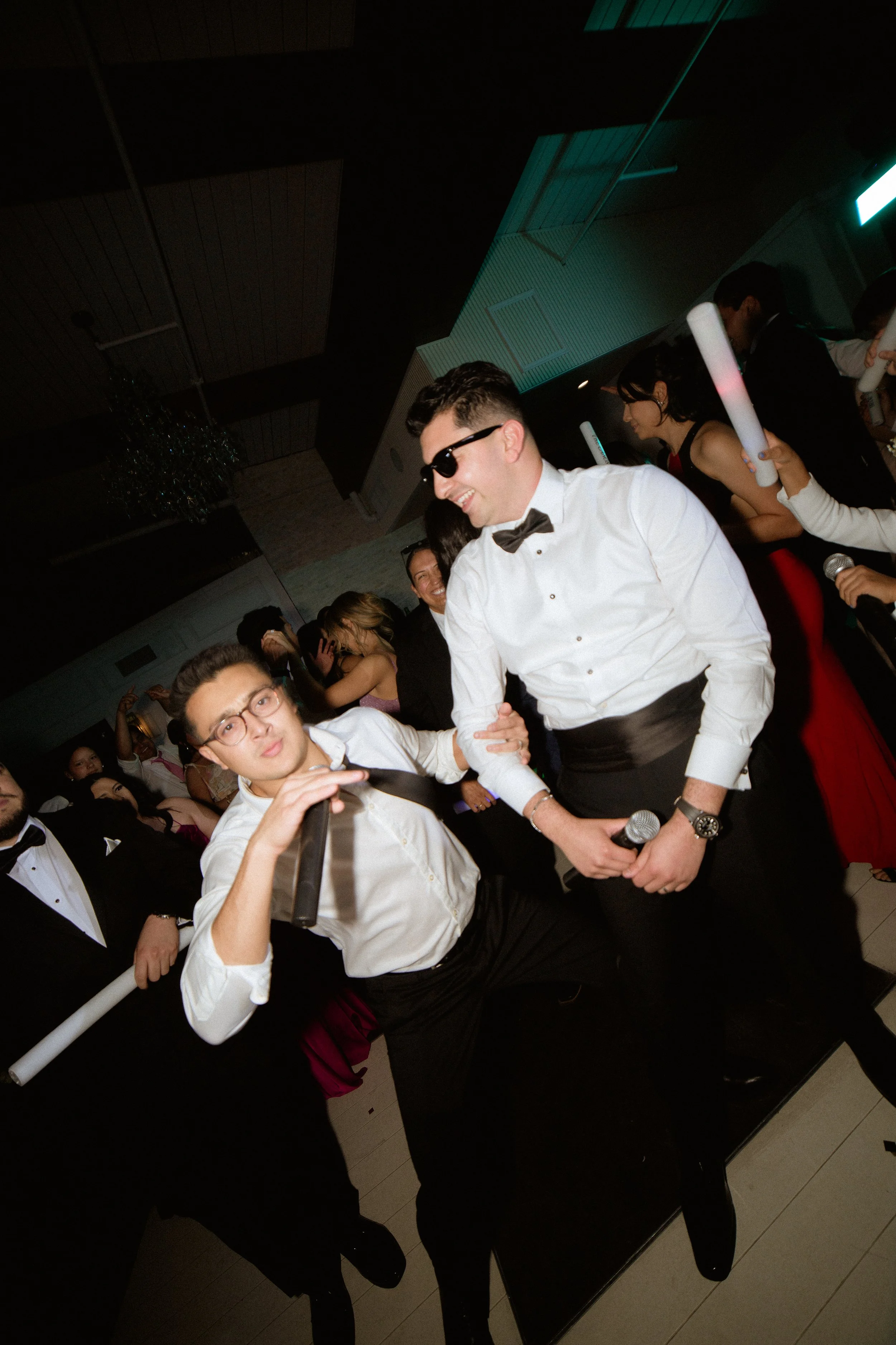 People dressed in formal attire at a celebration, with some holding rolled-up papers, indoors with dim lighting.