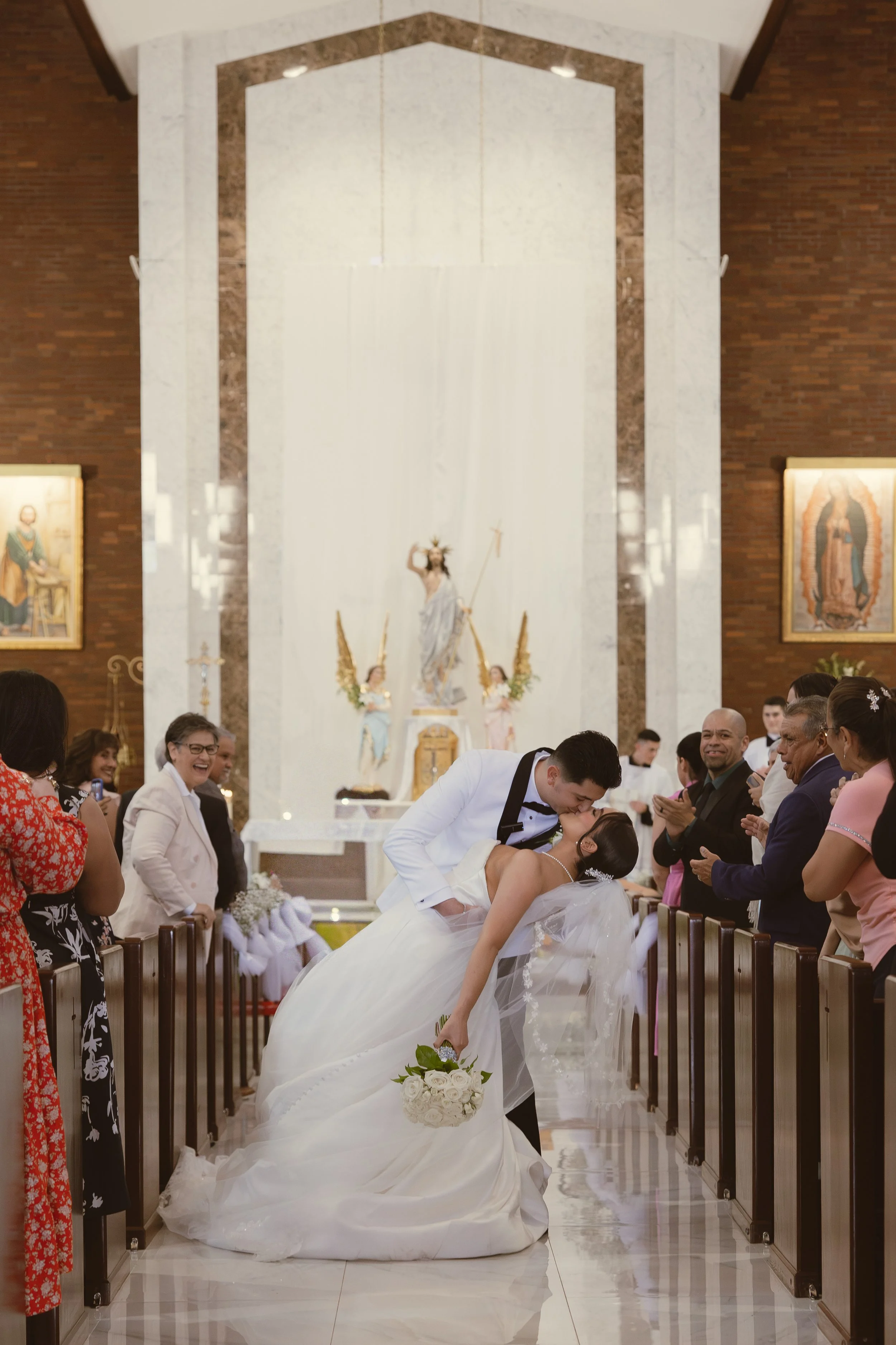 A wedding ceremony inside a church, with the bride and groom sharing a kiss. The bride is holding a bouquet of white roses, and there are guests smiling and clapping around them. Religious artwork decorates the church interior.