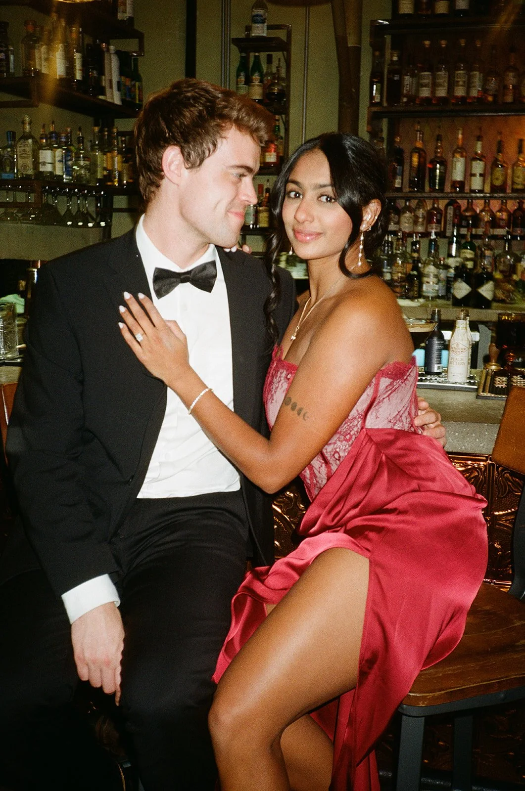 A man in a tuxedo and a woman in a red dress posing together in a bar or restaurant setting with shelves of liquor bottles in the background.
