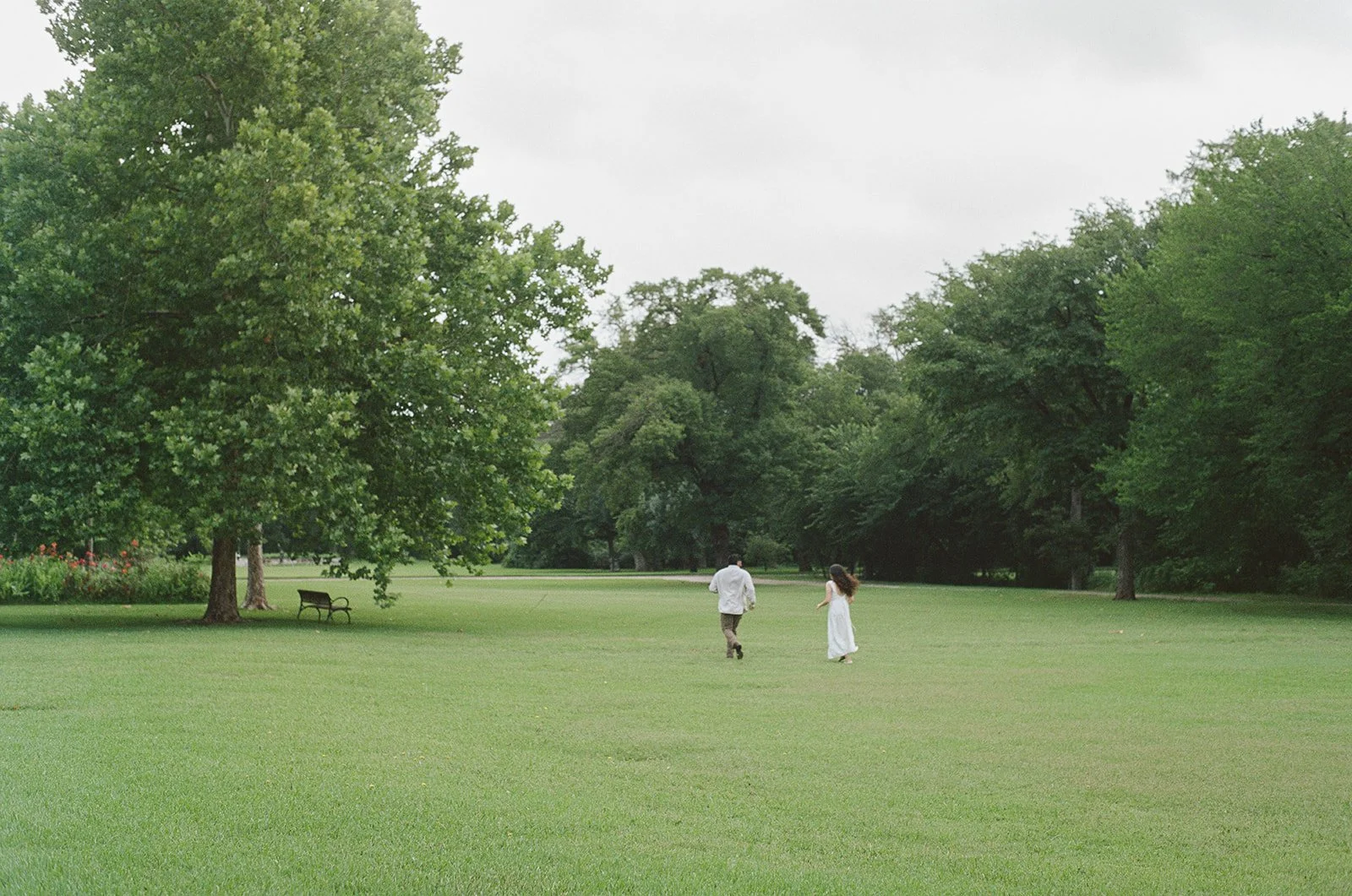 A couple walking in a lush green park with large trees, benches, and a cloudy sky.