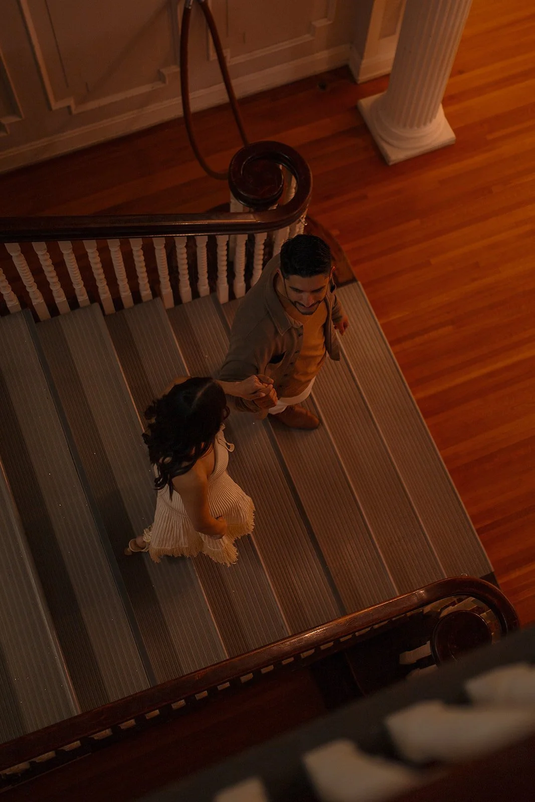 A man and woman holding hands while walking down a staircase in a warmly lit interior with wooden floors and a column.
