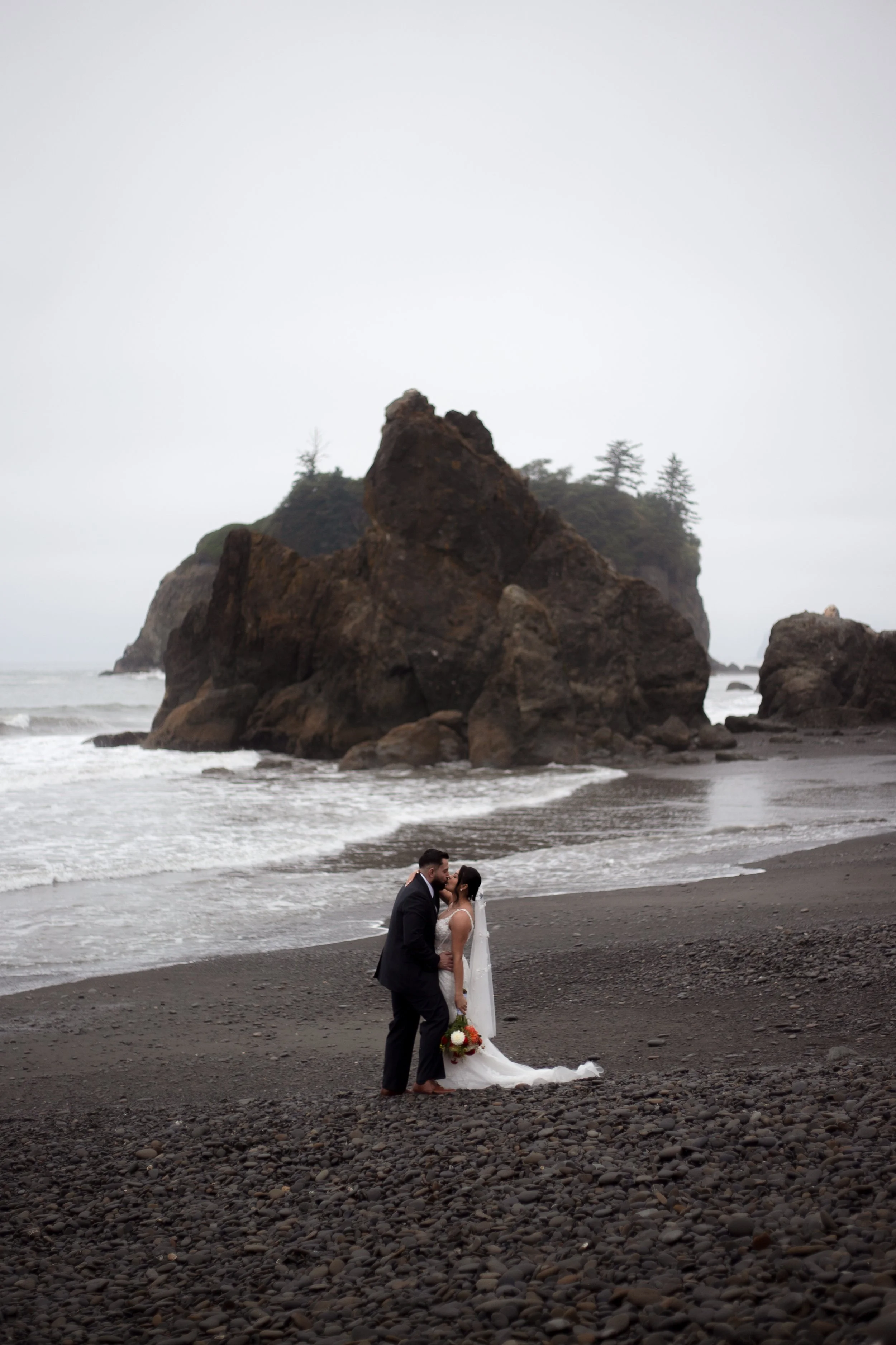 A bride and groom sharing a kiss on a rocky beach with large sea stacks and foggy sky in the background.