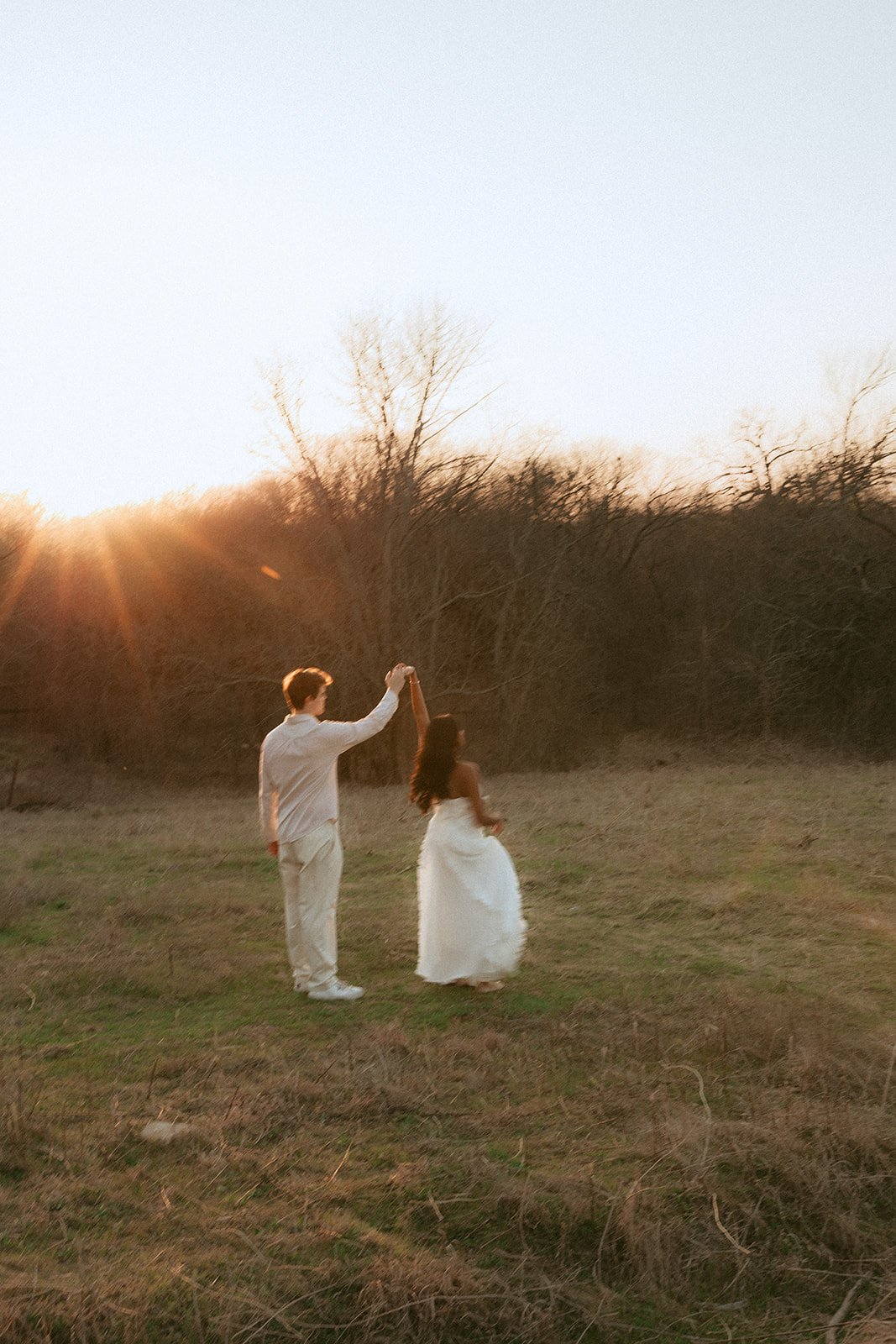A couple dancing outdoors at sunset; the man is holding the woman's hand as she twirls in a white dress with trees in the background.