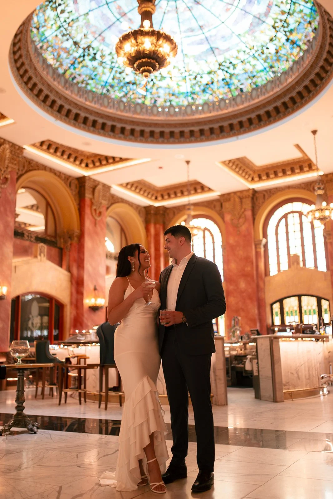 A couple in formal attire enjoying a drink and a chat inside an elegant, ornately decorated building with a stained glass dome ceiling and large arched windows.