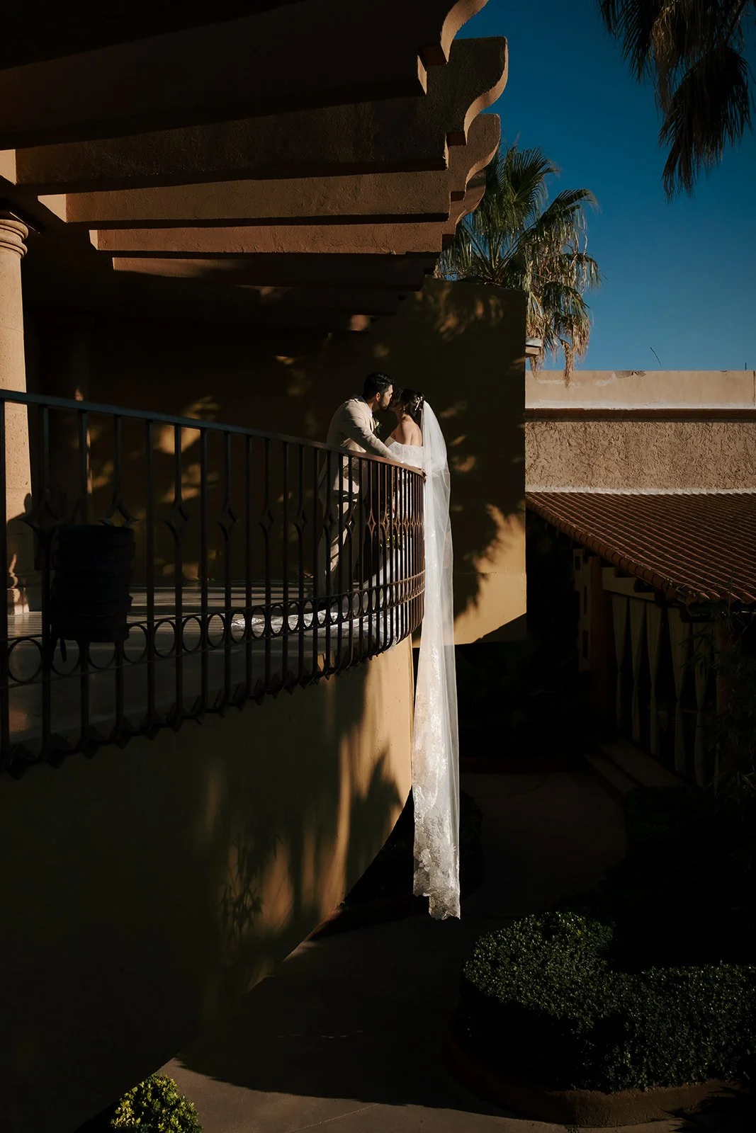A bride and groom stand on a curved balcony at sunset, sharing an intimate moment with the bride wearing a long veil and the groom in a light-colored suit. Shadows from nearby trees cast patterns on the wall, with blue sky and palm trees in the background.