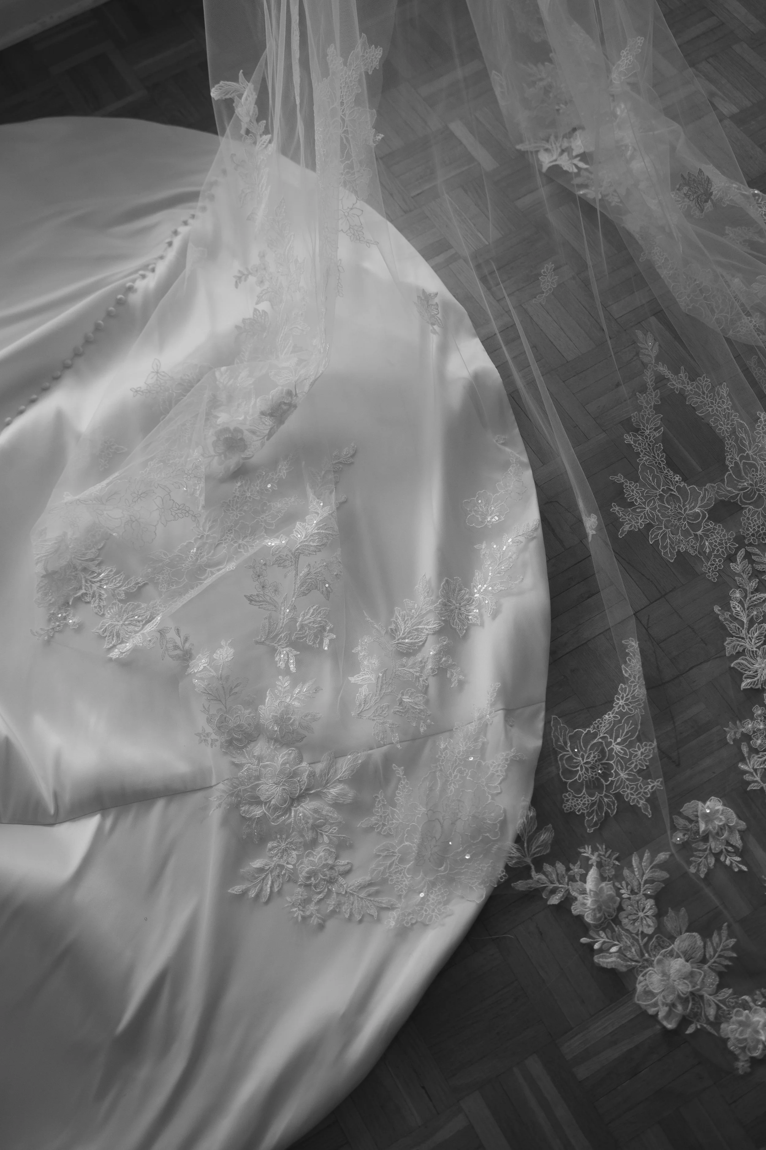 A wedding dress with intricate floral embroidery lying on a wooden floor, with a sheer lace veil featuring similar embroidery draped over it.