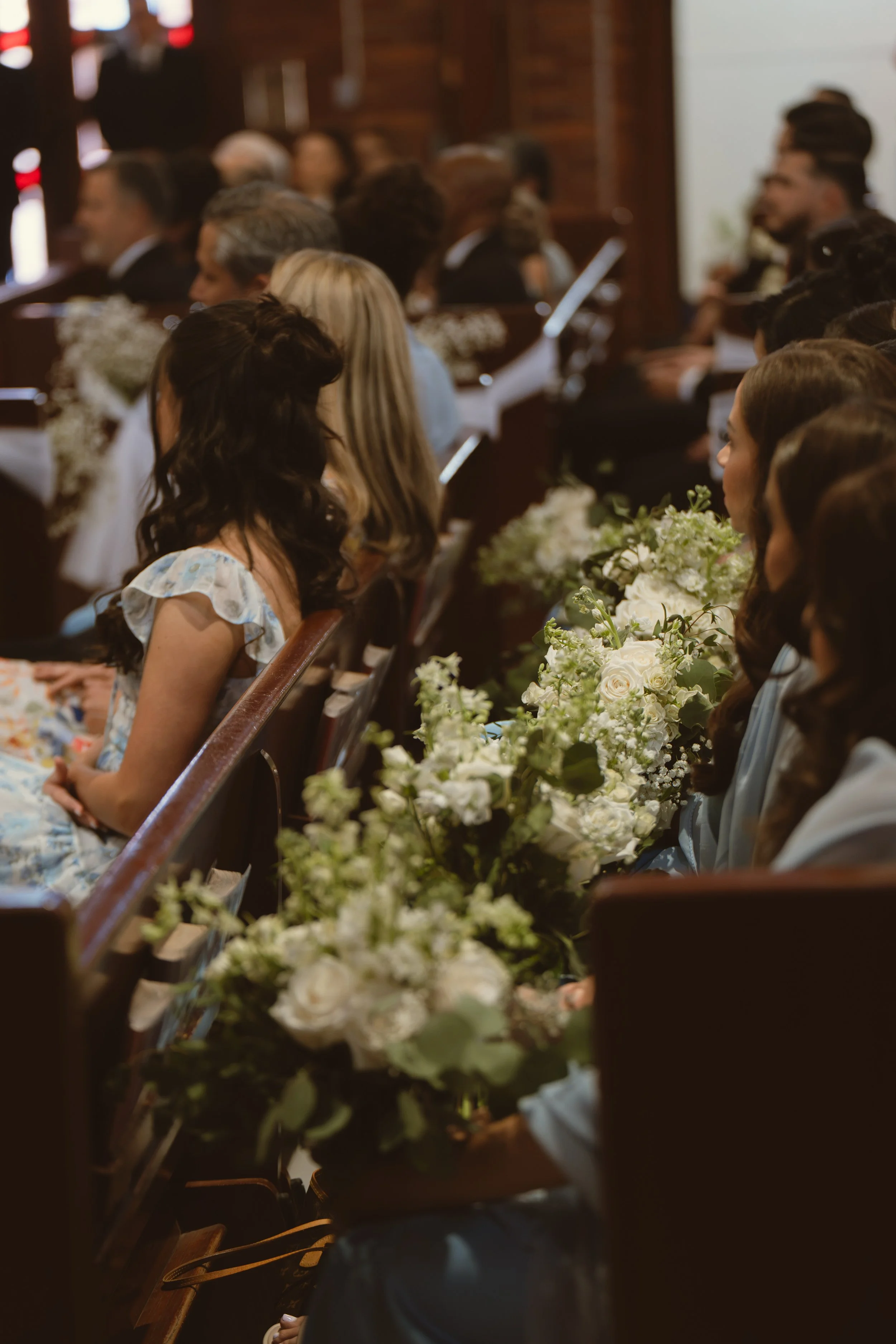 A group of people seated inside a church or chapel, attending a ceremony, with floral arrangements on their tables.