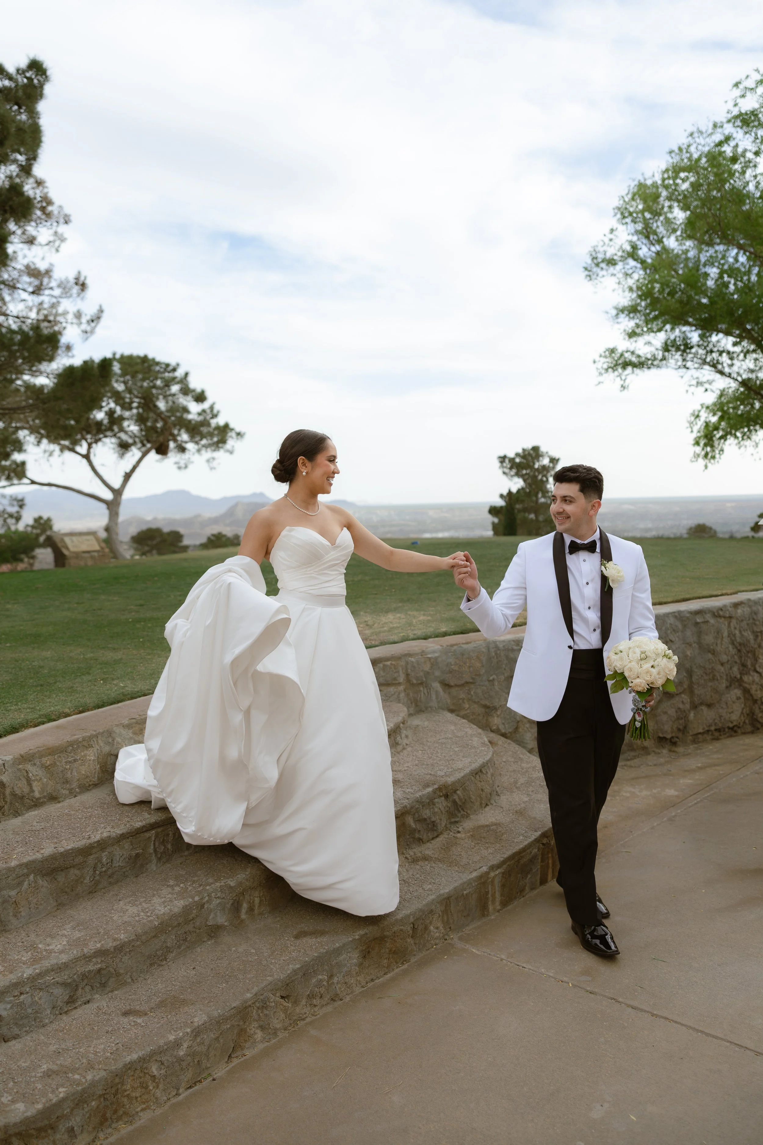A newlywed couple holding hands in an outdoor park setting with trees and hills in the background. The bride is wearing a strapless white wedding gown and the groom is dressed in a tuxedo with a bow tie and holding a bouquet of white roses.