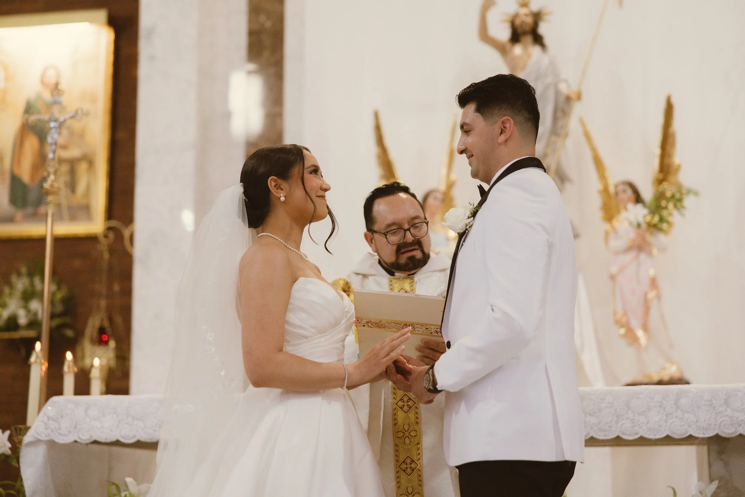 Bride and groom exchanging vows at a church wedding ceremony, with a priest officiating, in front of religious icons and decorative candles.