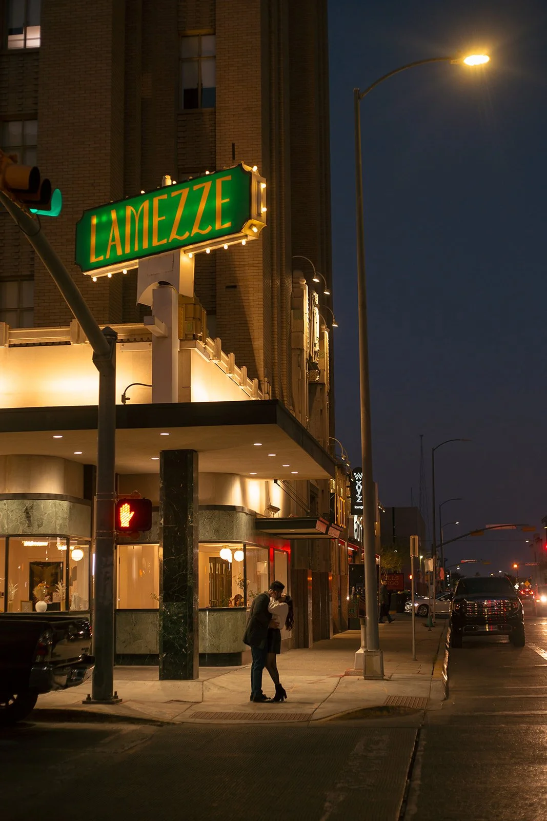 Nighttime street scene with a couple kissing on the sidewalk, illuminated by streetlights and building lights. A large green sign reads 'LAMEZZE' in yellow letters. Cars are parked along the street, and a traffic light shows a red hand signal for ped