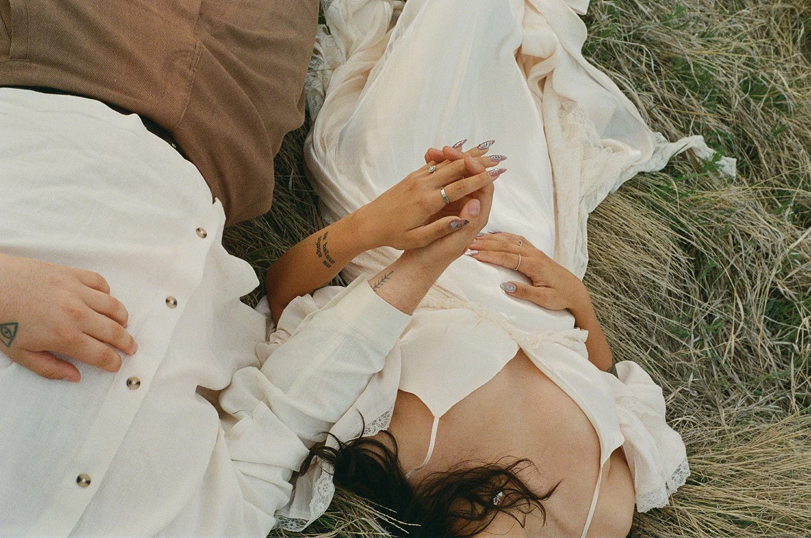 Two people lying on grass, one wearing a white blouse and the other in brown pants. The person in white has long dark hair and rings on fingers. The other has their hand on the white blouse, showing a tattoo and jewelry. Their faces are not visible.