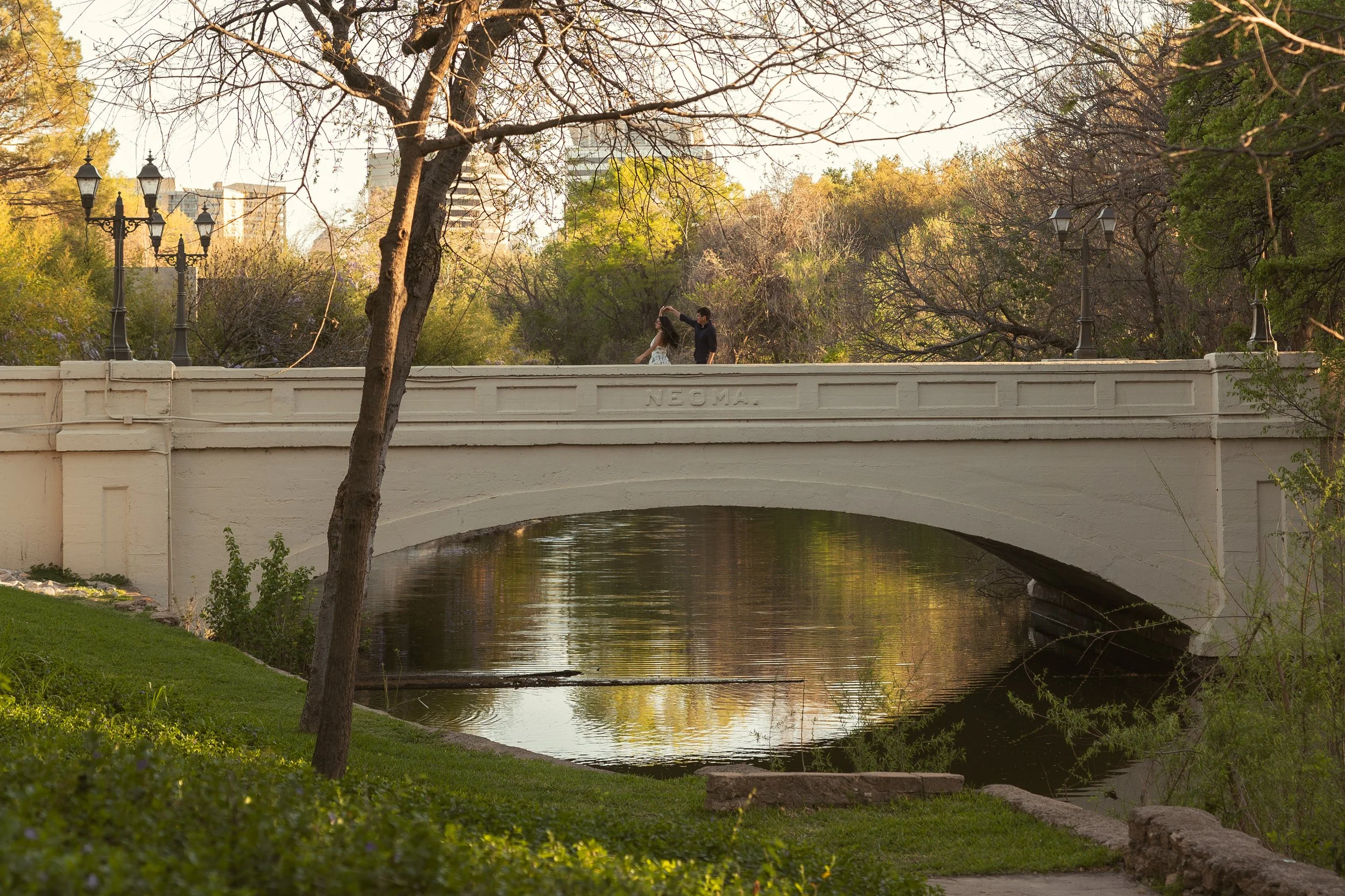 A bridge over a small river in a park with two people dancing and a tree with bare branches in the foreground.