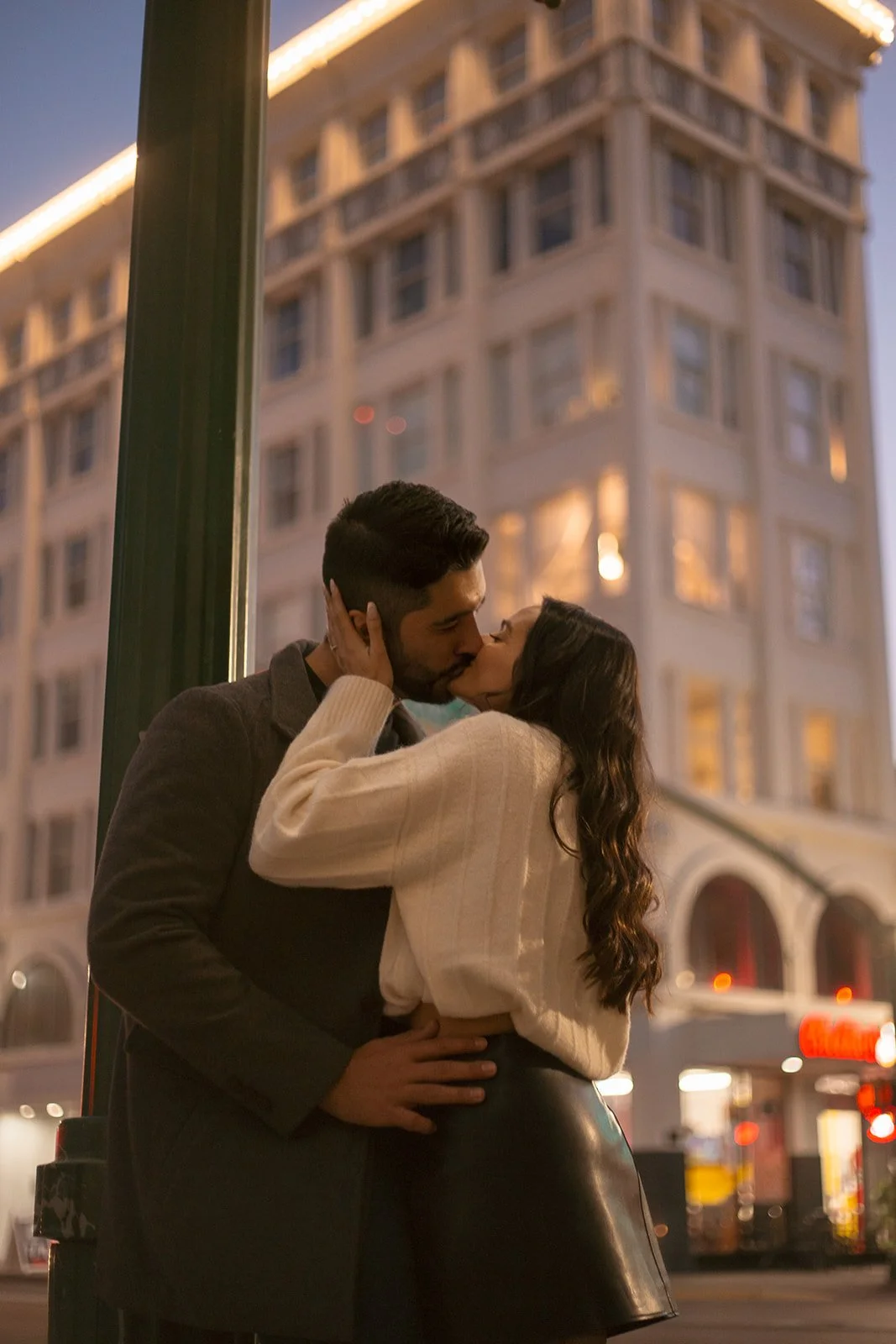 A couple is kissing on a city street at dusk, with tall buildings and warm city lights in the background.