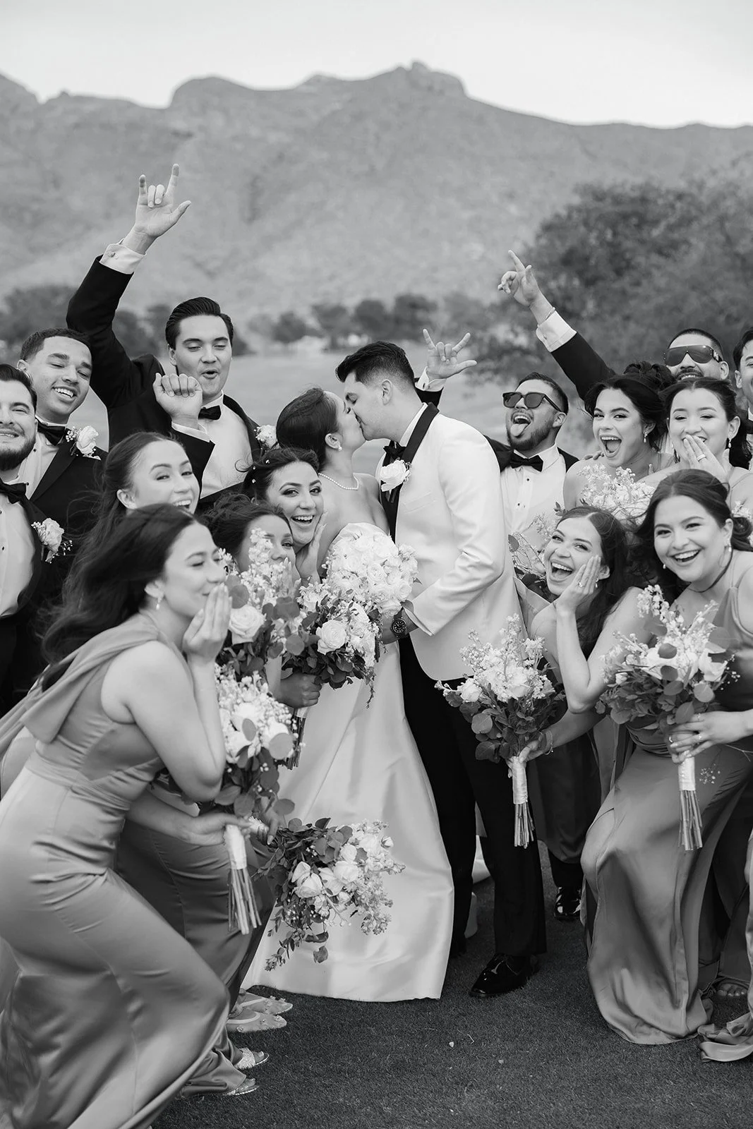 Black and white photo of a large wedding party celebrating outdoors. The bride and groom are kissing in the center, surrounded by their wedding guests who are smiling, laughing, and holding bouquets, with a mountain landscape in the background.