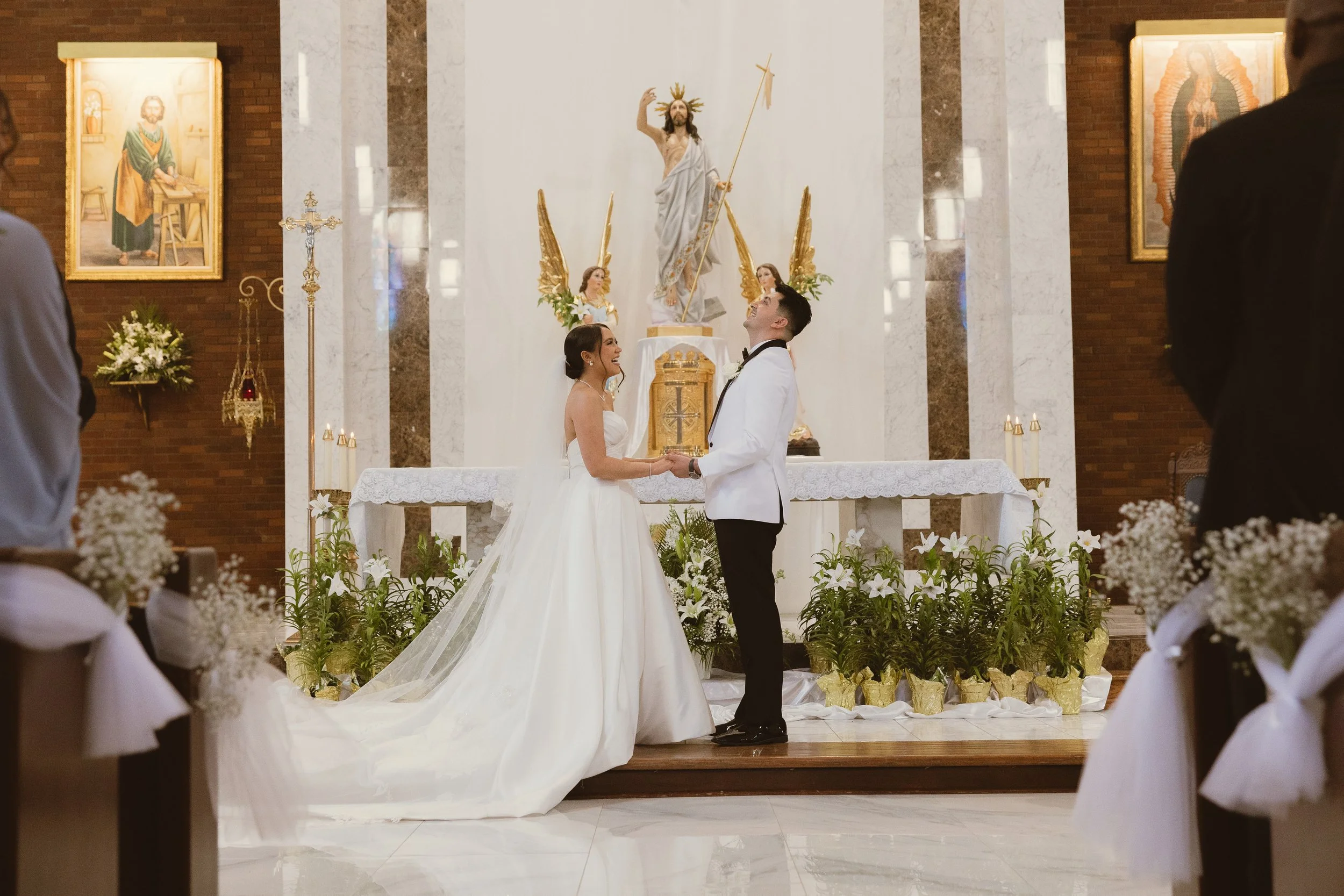 A bride and groom holding hands and smiling during their wedding ceremony inside a church, standing in front of an altar with religious artwork and statues.