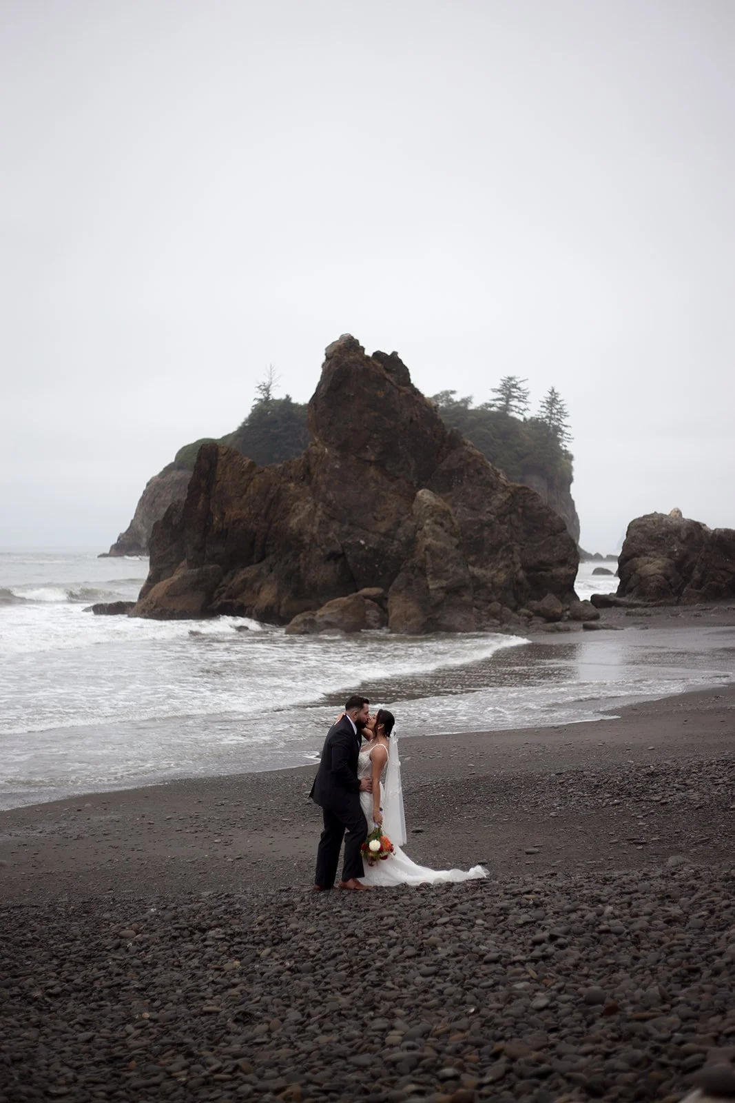 A bride and groom kissing on a dark pebble beach with large rock formations and an overcast sky in the background.