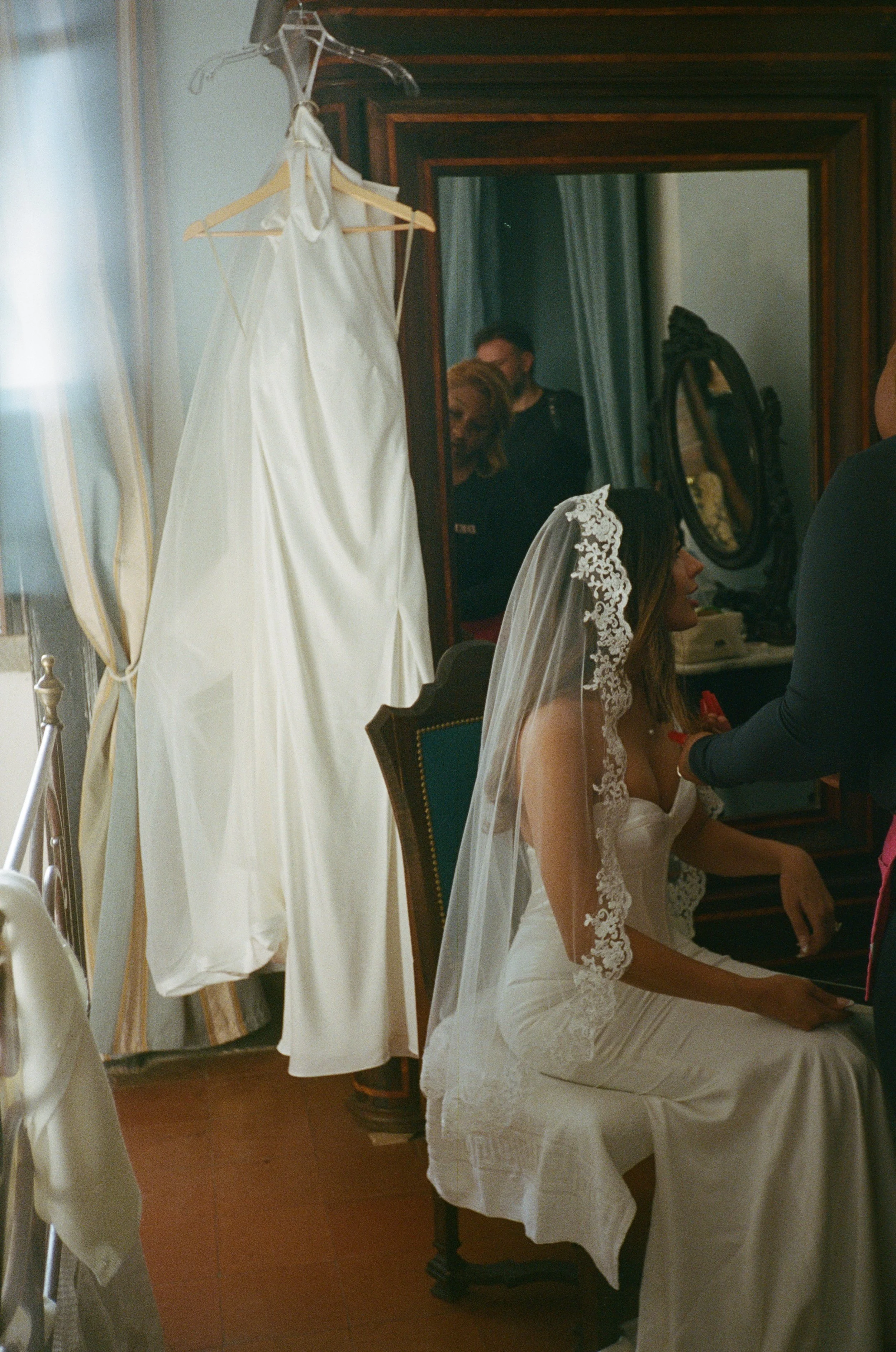 A woman sitting in a chair while a person adjusts her wedding dress in front of a mirror. Wedding dresses hanging on a clothing rack are visible in the foreground.