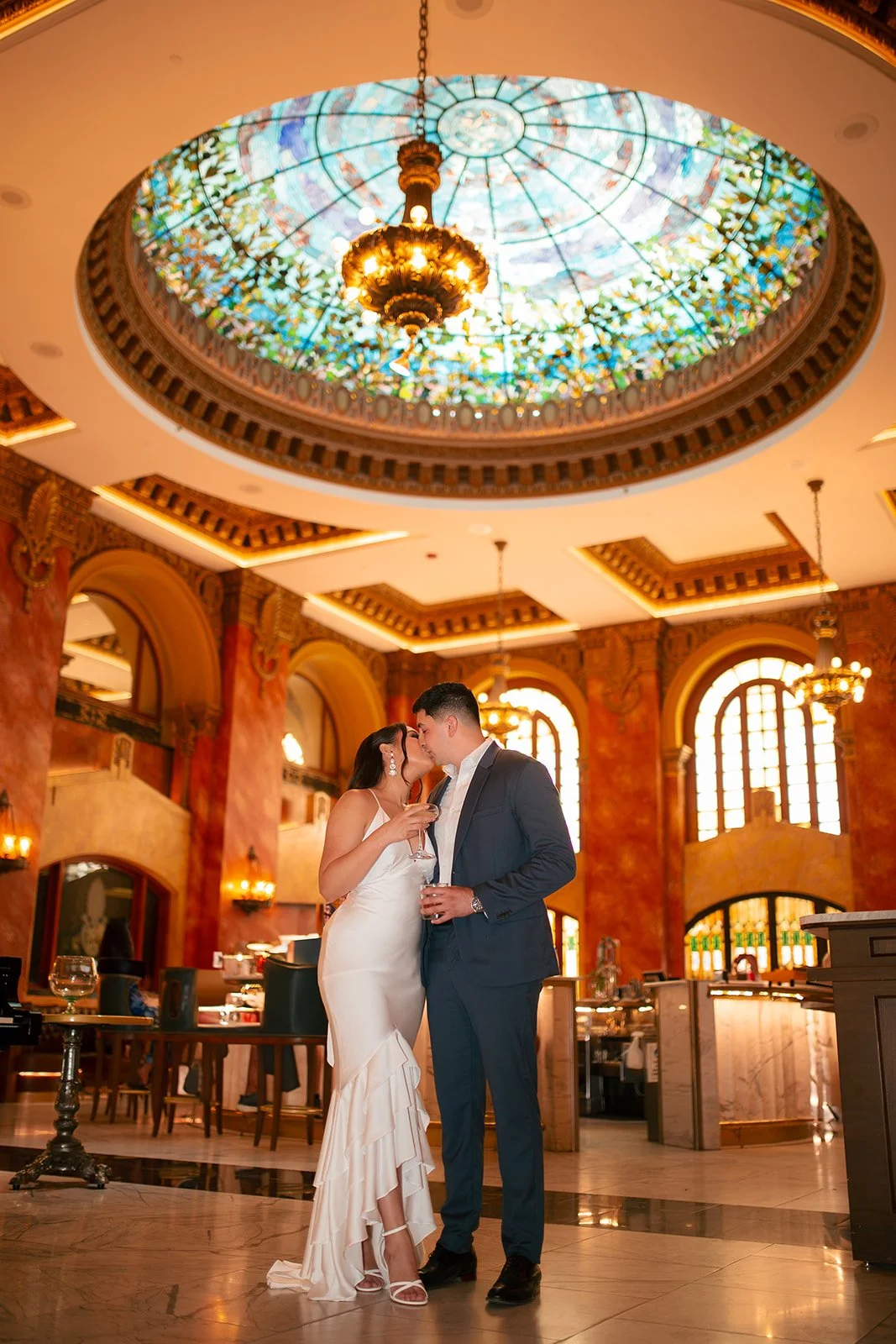 A couple, dressed in formal wedding attire, sharing a kiss inside an elegant, grand hall with a stained glass ceiling and ornate lighting fixtures.