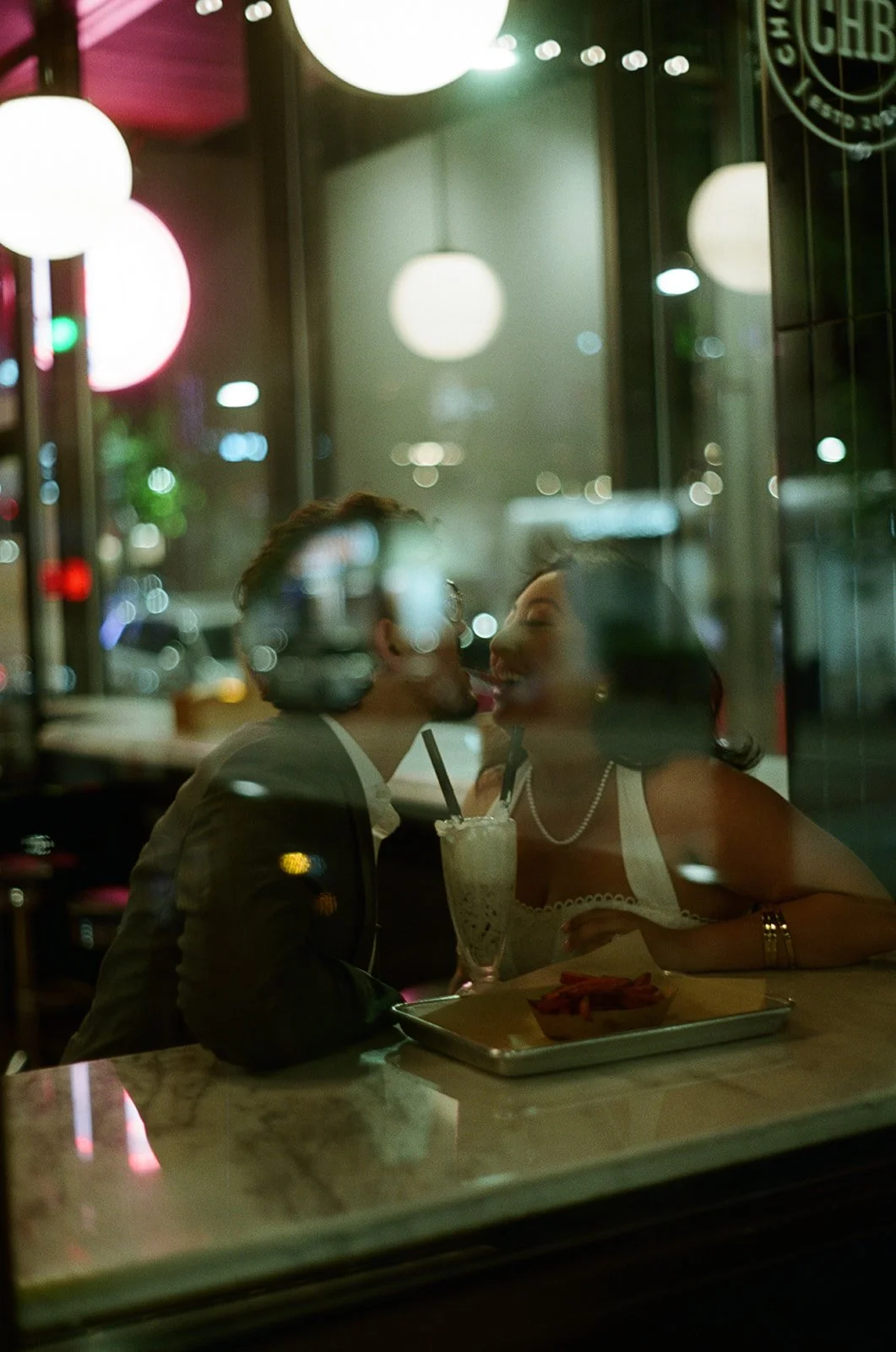 A romantic couple sharing a kiss through a restaurant window at night, with reflections of city lights and warm interior lighting.