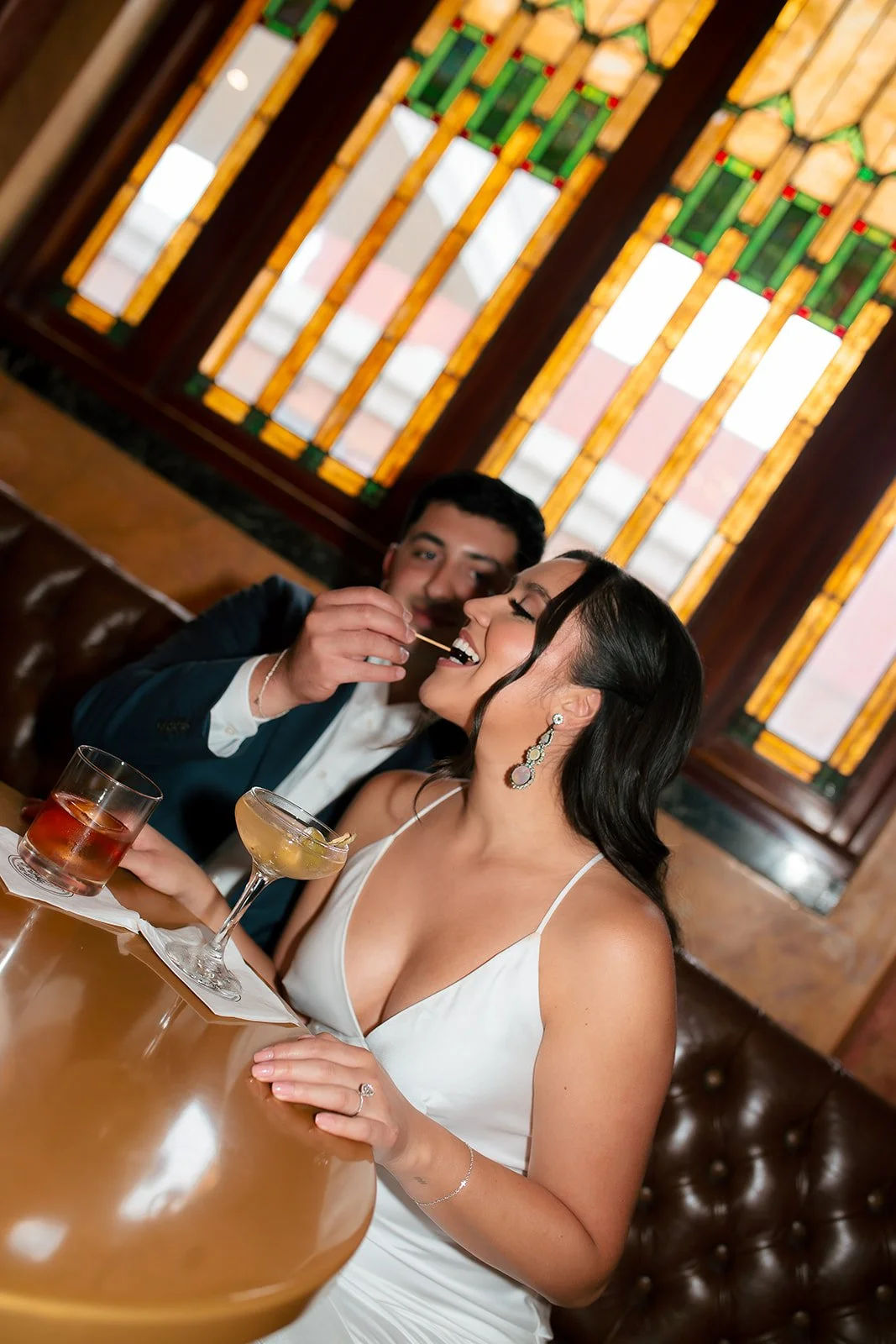 A woman in a white dress smiling while a man in a tuxedo feeds her a small piece of food at a restaurant table, with cocktails on the table and stained glass windows in the background.