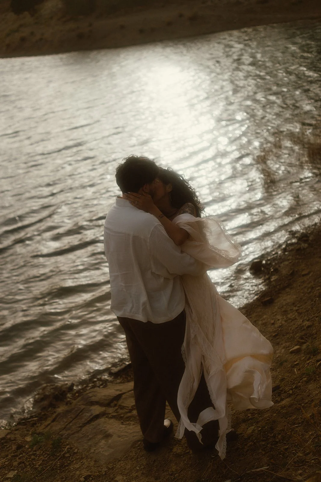 A couple embraces and kisses by a lake at sunset, with the sun reflecting on the water.