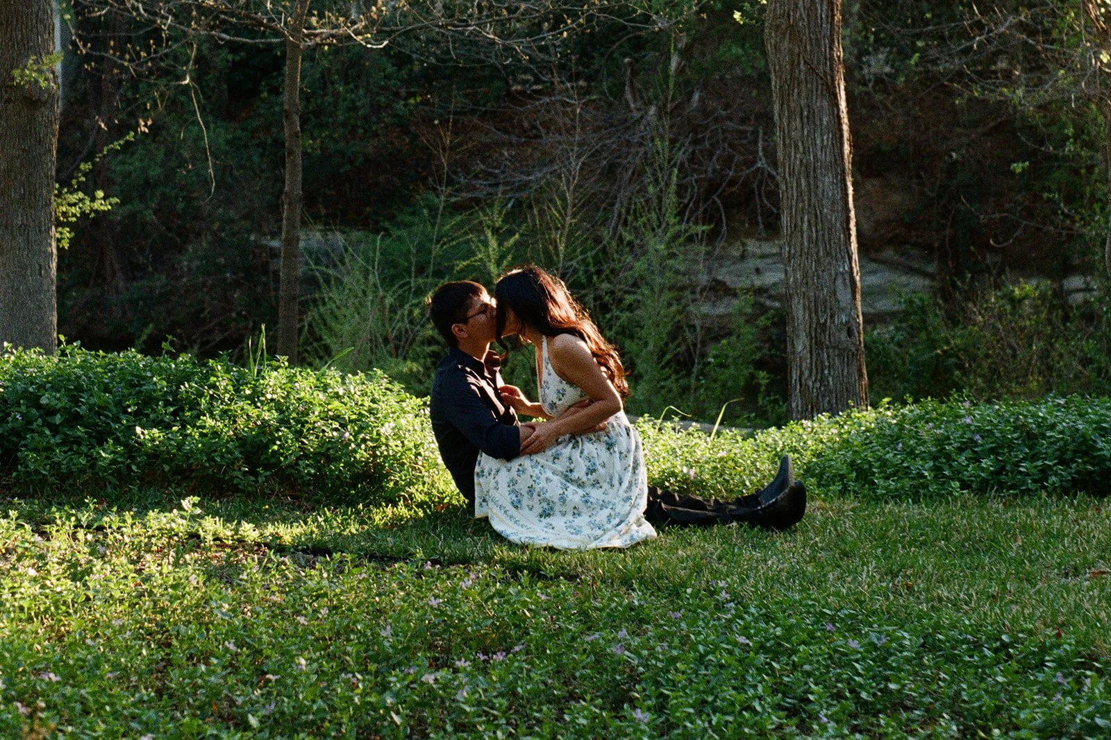 A couple kissing outdoors in a lush green park or garden with tall trees and bushes, sitting on the grass.