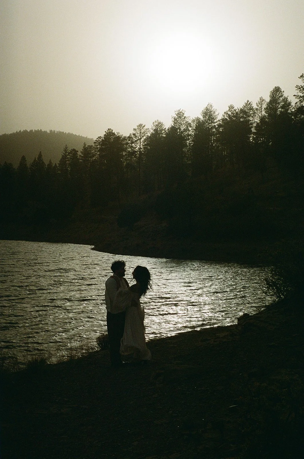 Silhouetted couple standing by a river at sunset with trees and hills in the background.