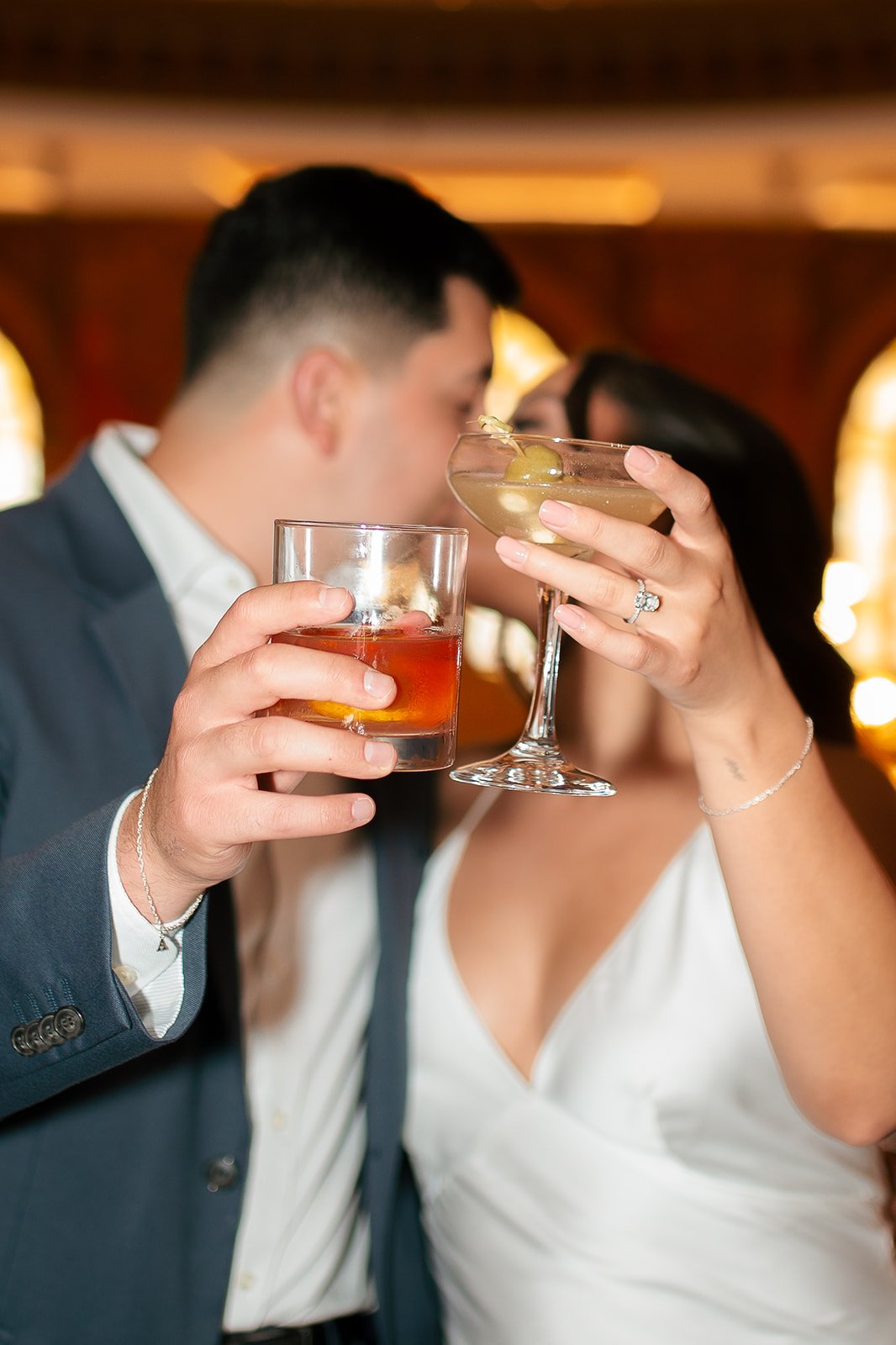 A couple sharing a romantic moment, holding drinks close to each other, with a blurred background and warm lighting.